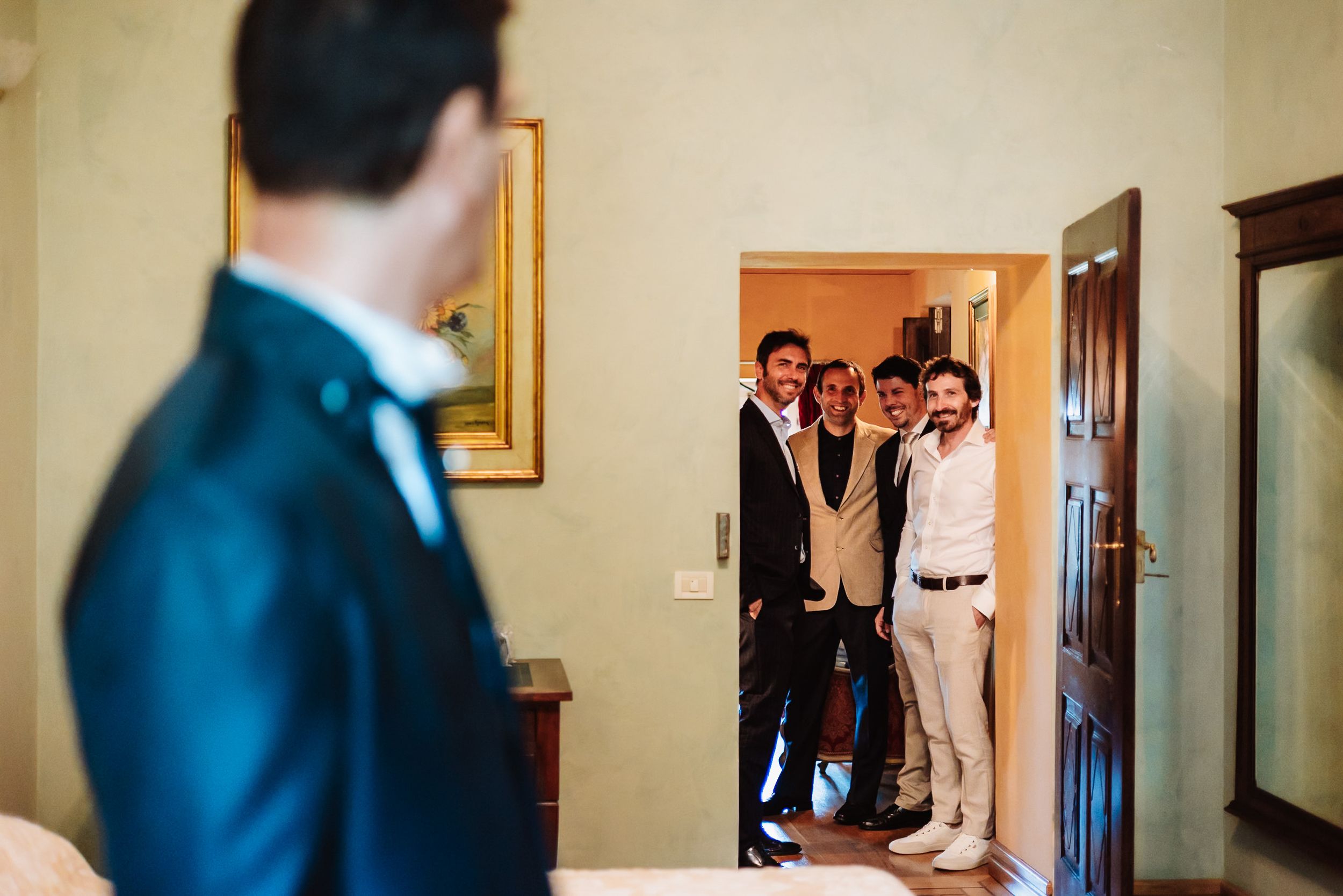 Groom in navy suit sees groomsmen smiling in doorway during wedding morning preparation.