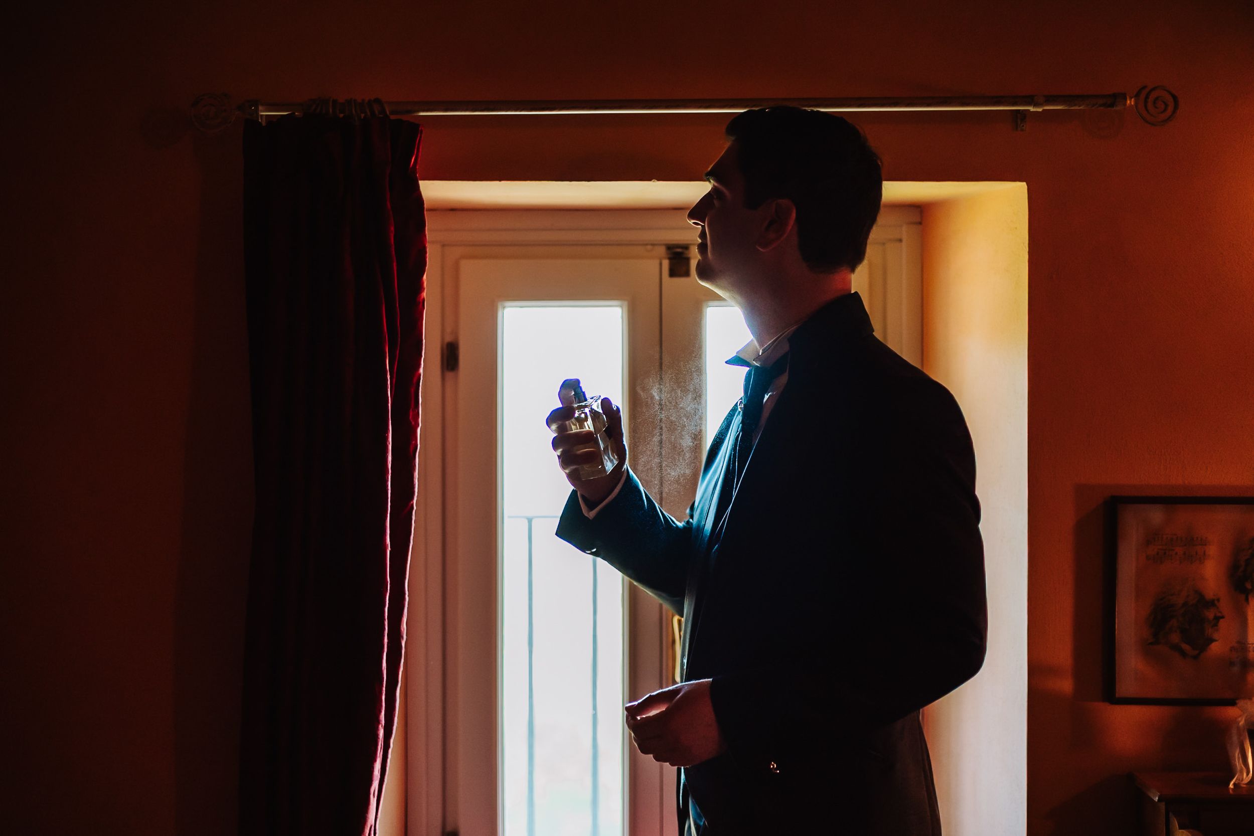 Groom in dark suit applying cologne by window in warm-lit room before wedding ceremony.
