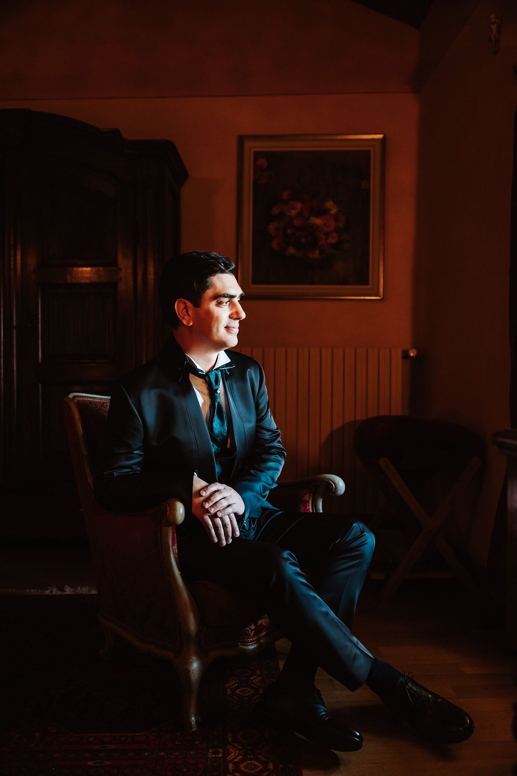 Elegant man in dark suit sitting in vintage chair, gazing sideways in warmly lit room with classic painting.