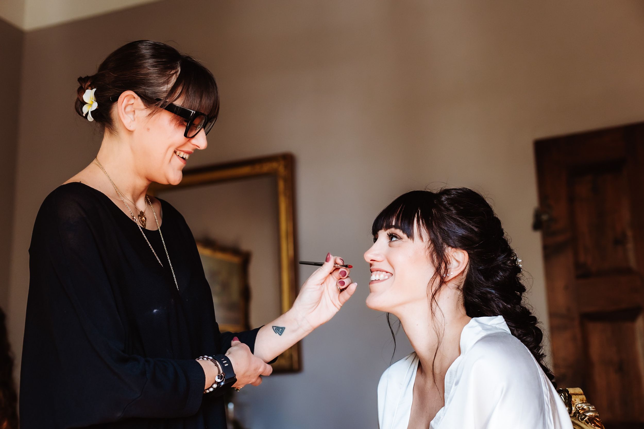 Makeup artist applying makeup to a smiling bride in a white robe during wedding preparations.