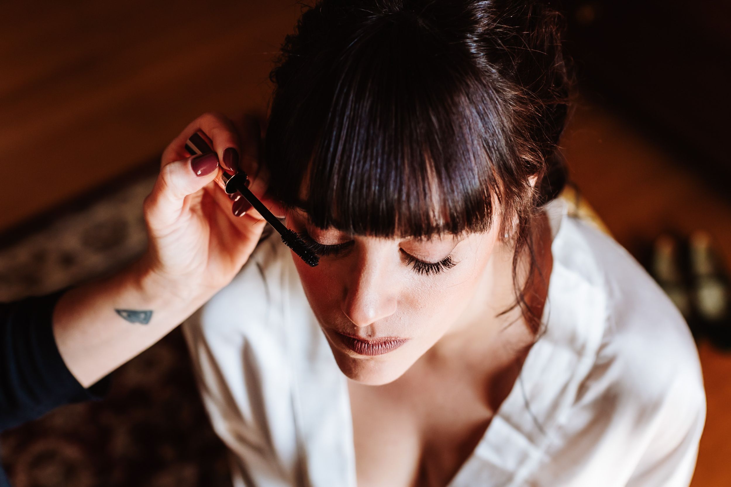 Bride getting mascara applied during wedding morning preparations, wearing white robe with dark updo hairstyle.