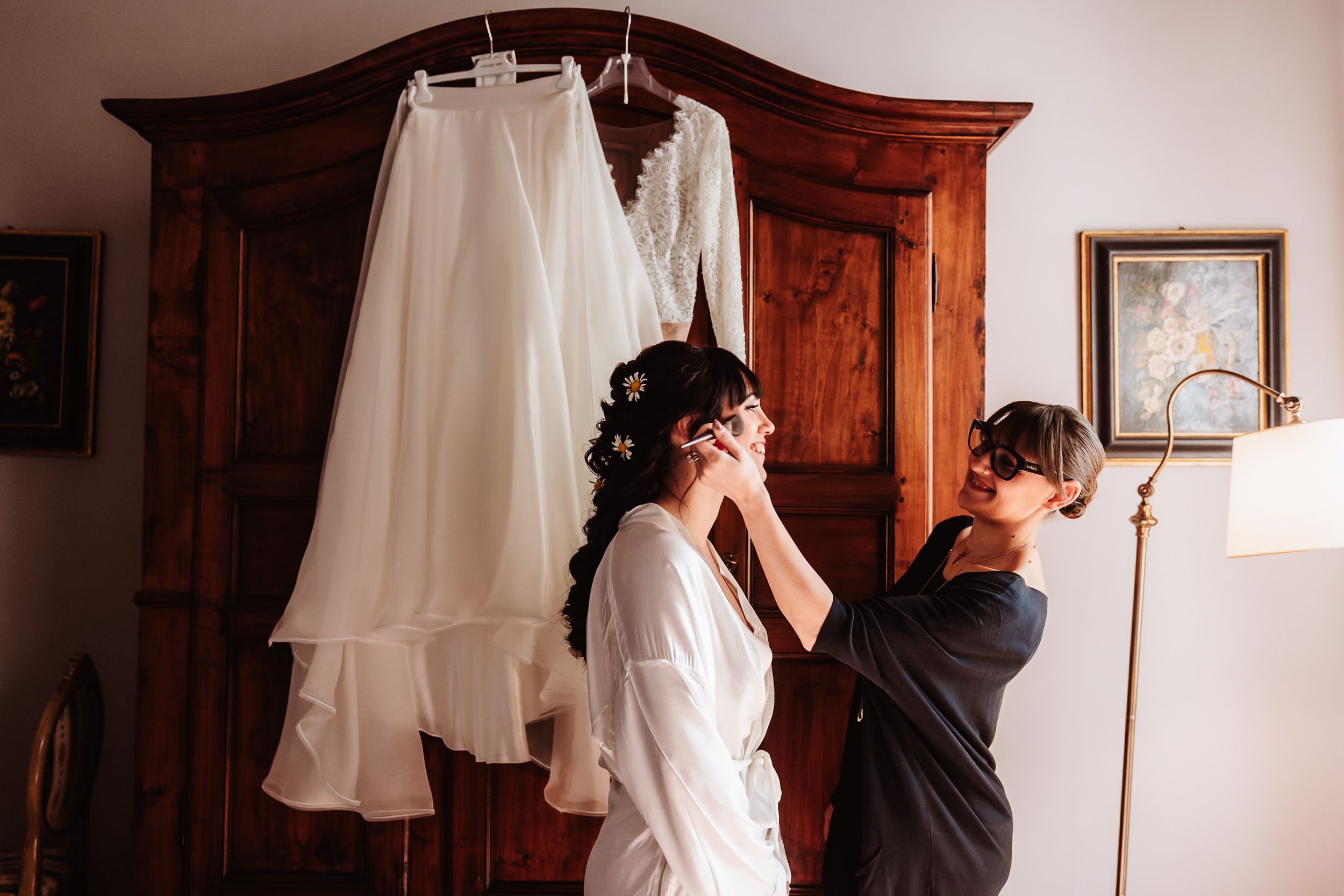 Bride in white robe getting earrings put on near wooden armoire with wedding dress hanging above.
