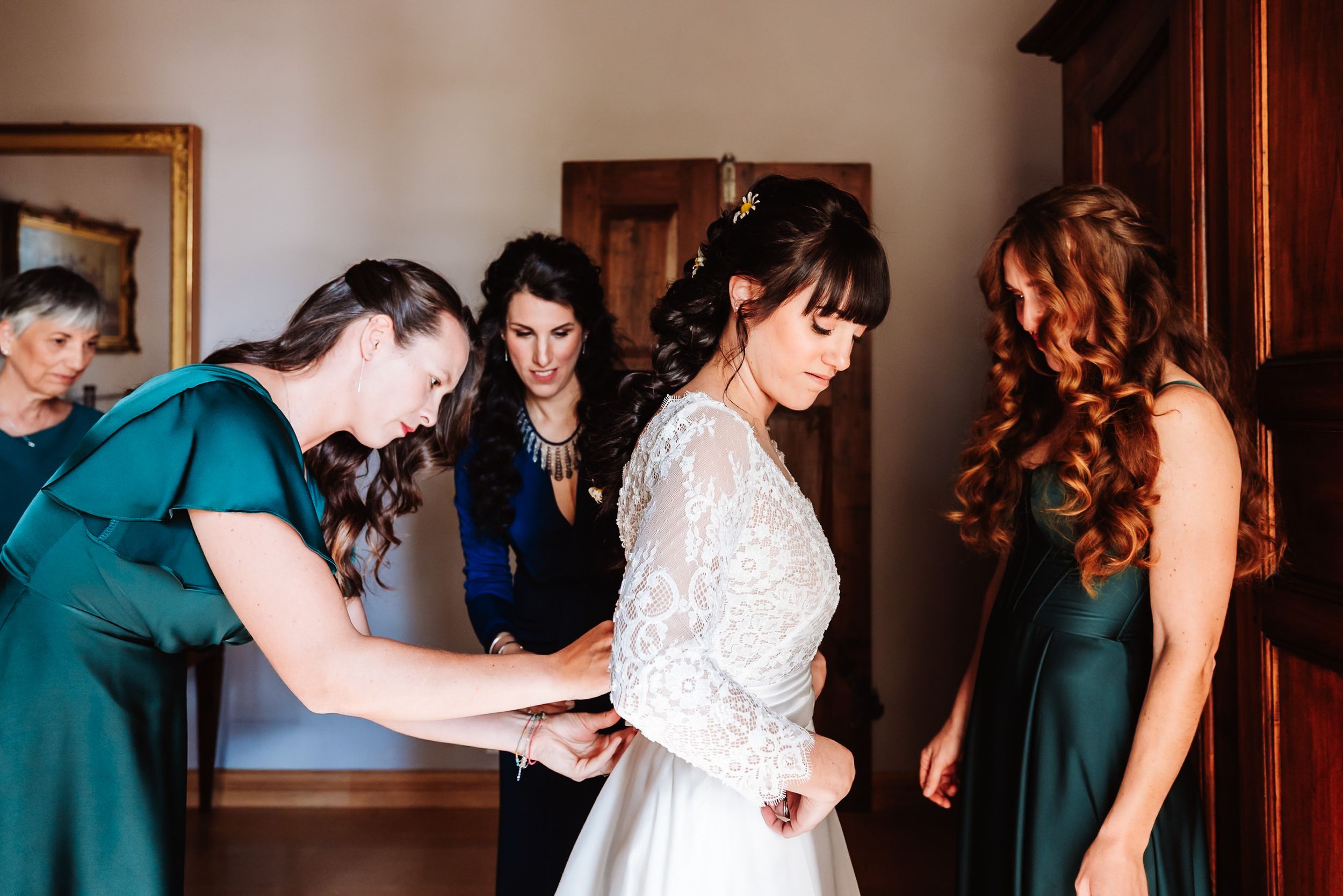 Bridesmaids in emerald green dresses helping bride with wedding gown preparations in elegant room.