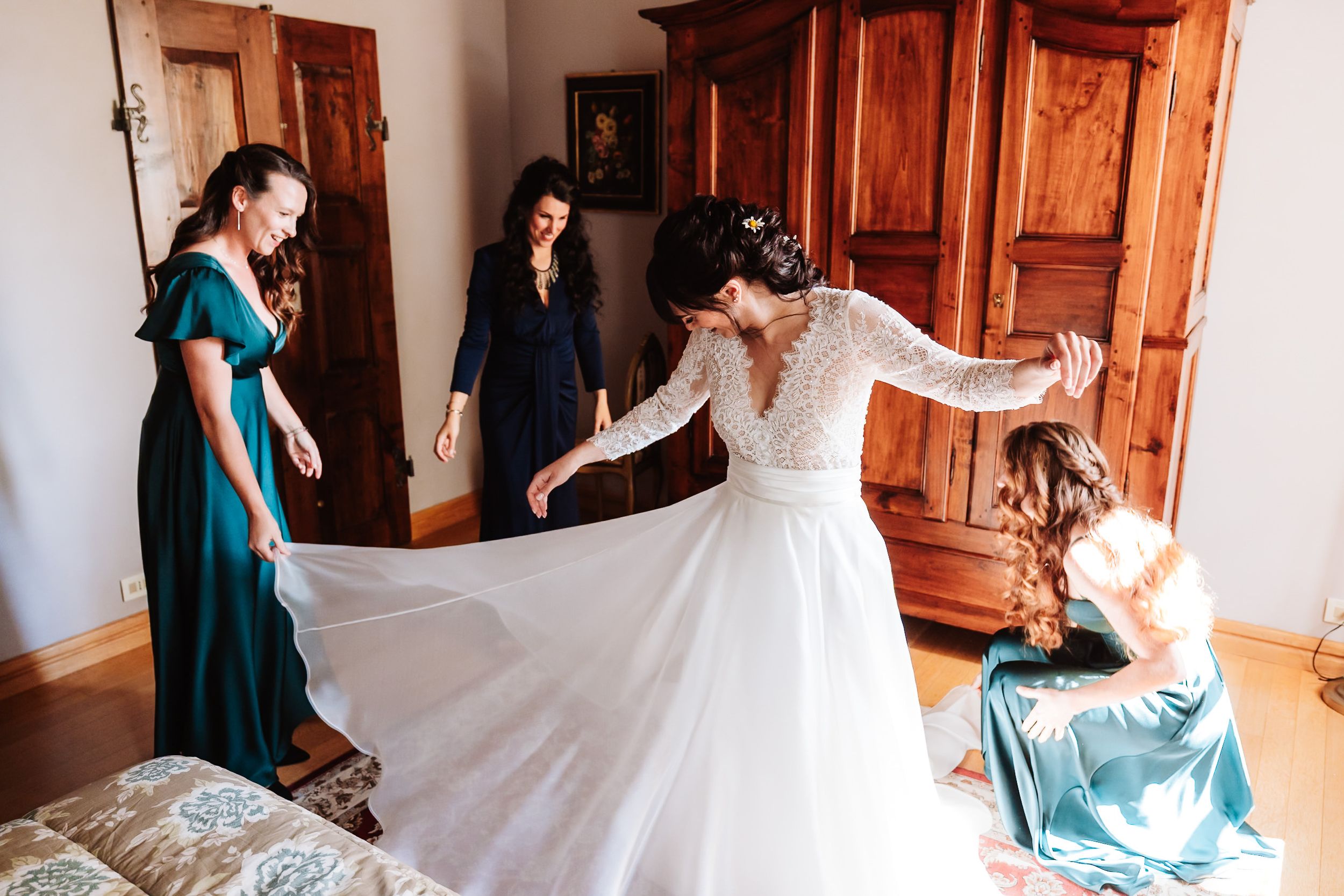 Bride in lace wedding gown twirling skirt assisted by bridesmaids in teal and blue dresses.