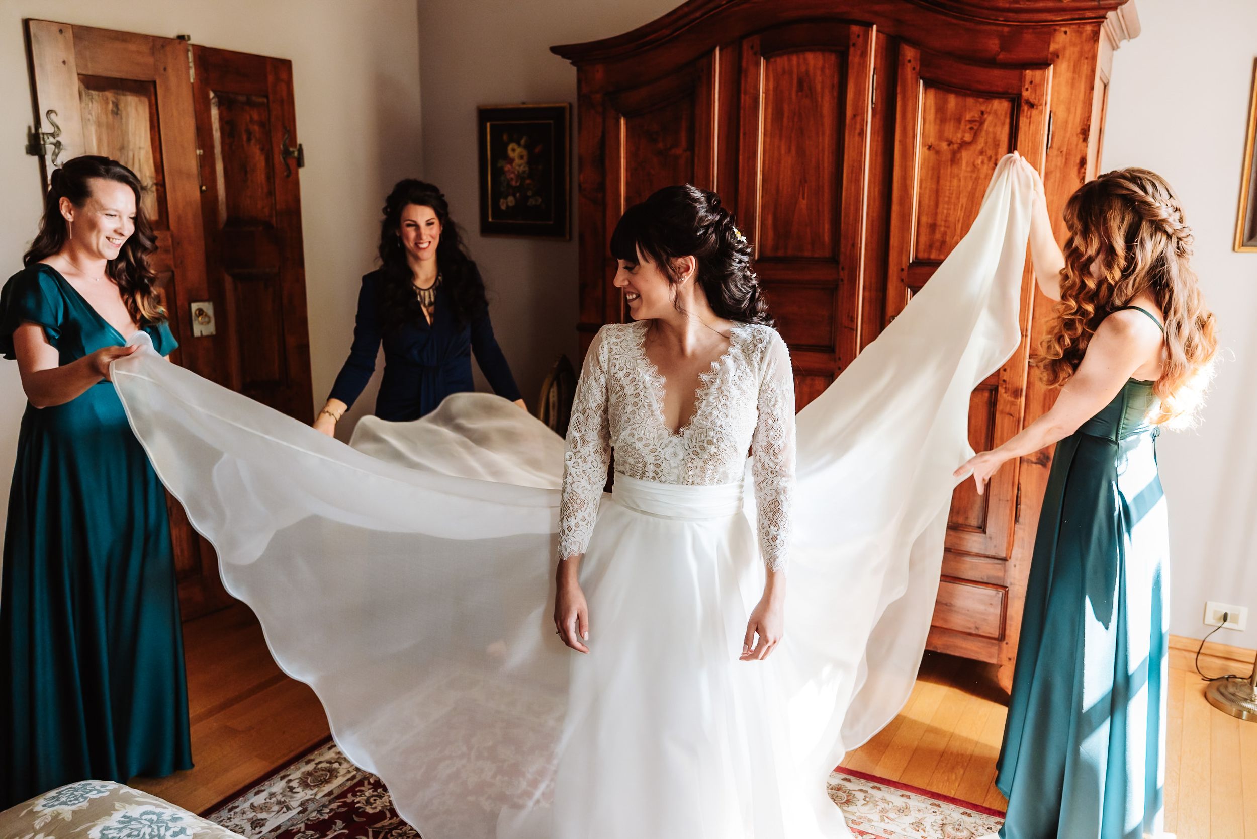 Bridesmaids spreading bride's white wedding veil in elegant room with wooden armoire.