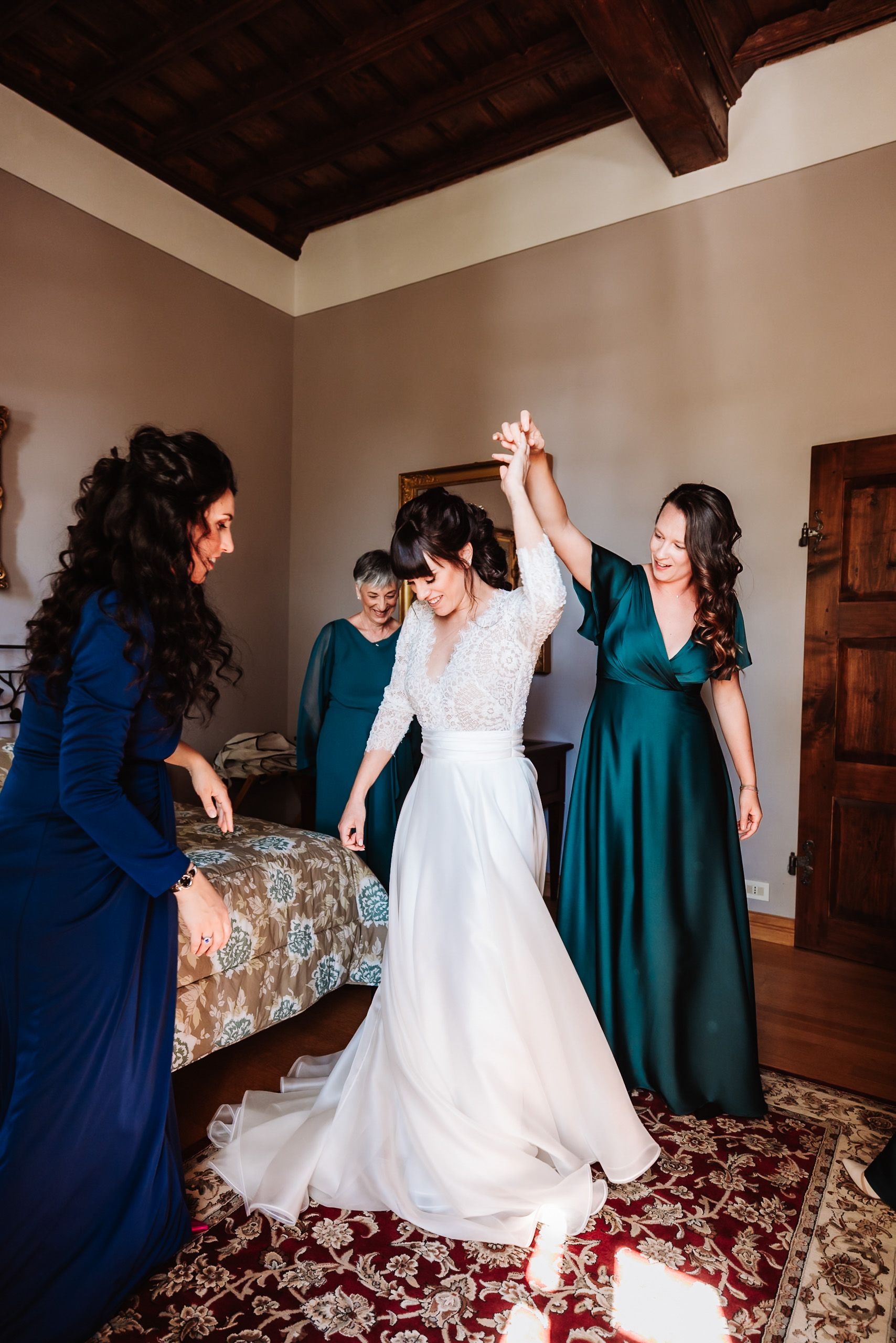 Bride twirling in white gown assisted by bridesmaids in blue and green dresses in elegant room.