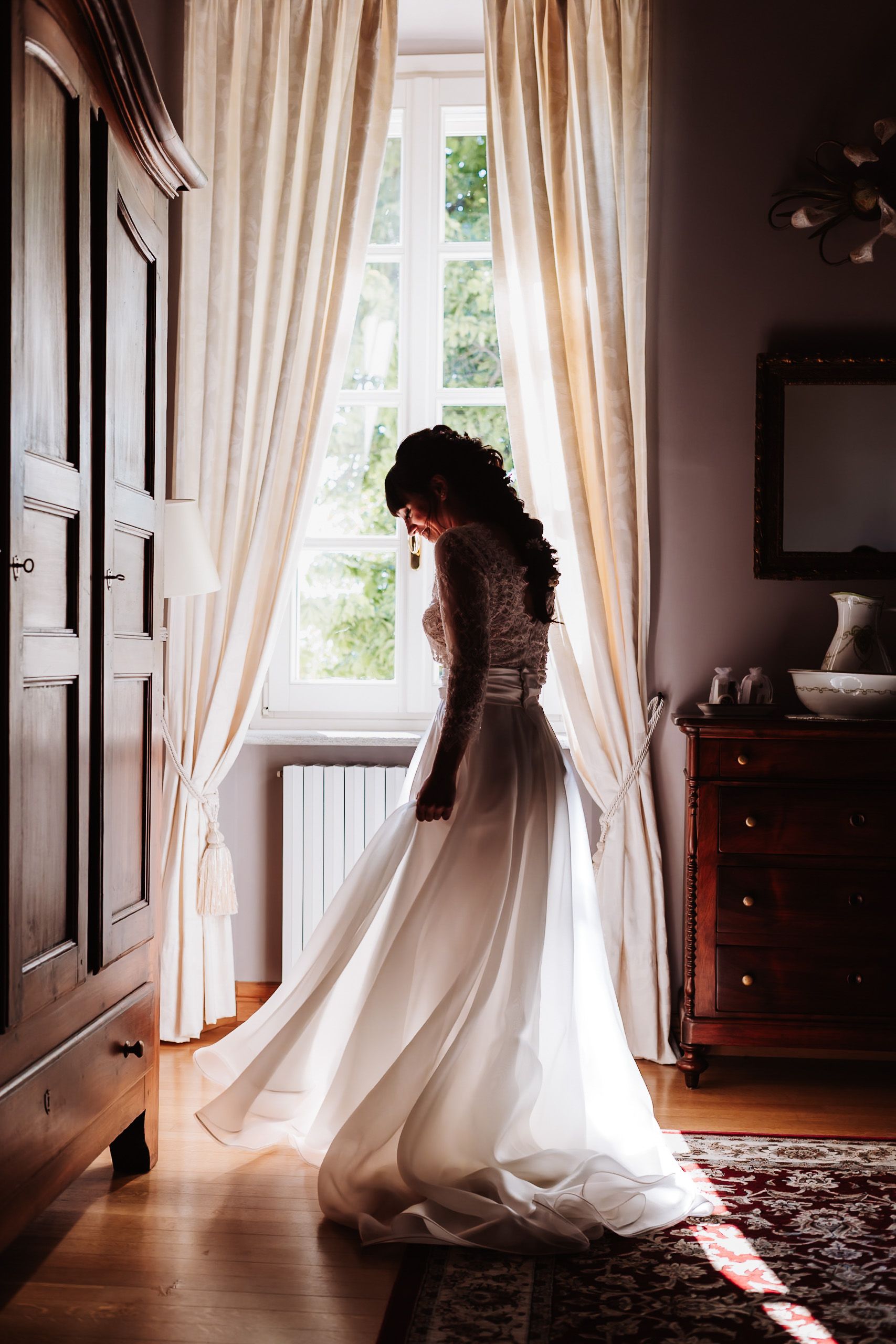 Bride in elegant white wedding gown standing by sunlit window with flowing curtains in a classic interior room.