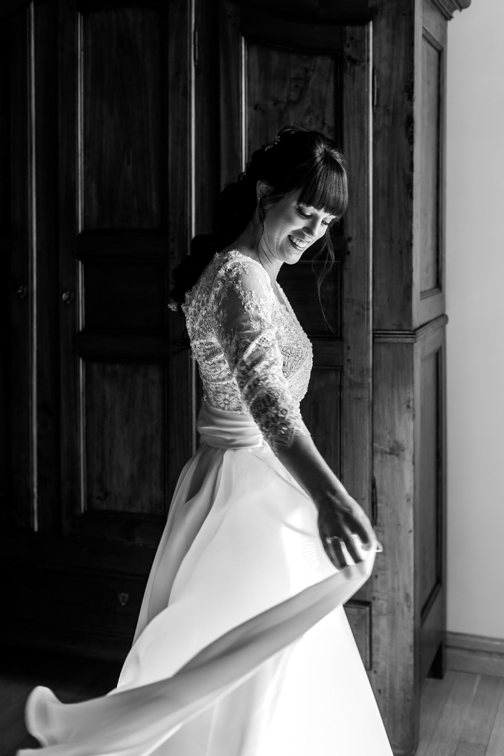 Bride in lace wedding dress twirling joyfully by wooden doors in elegant black and white photo.