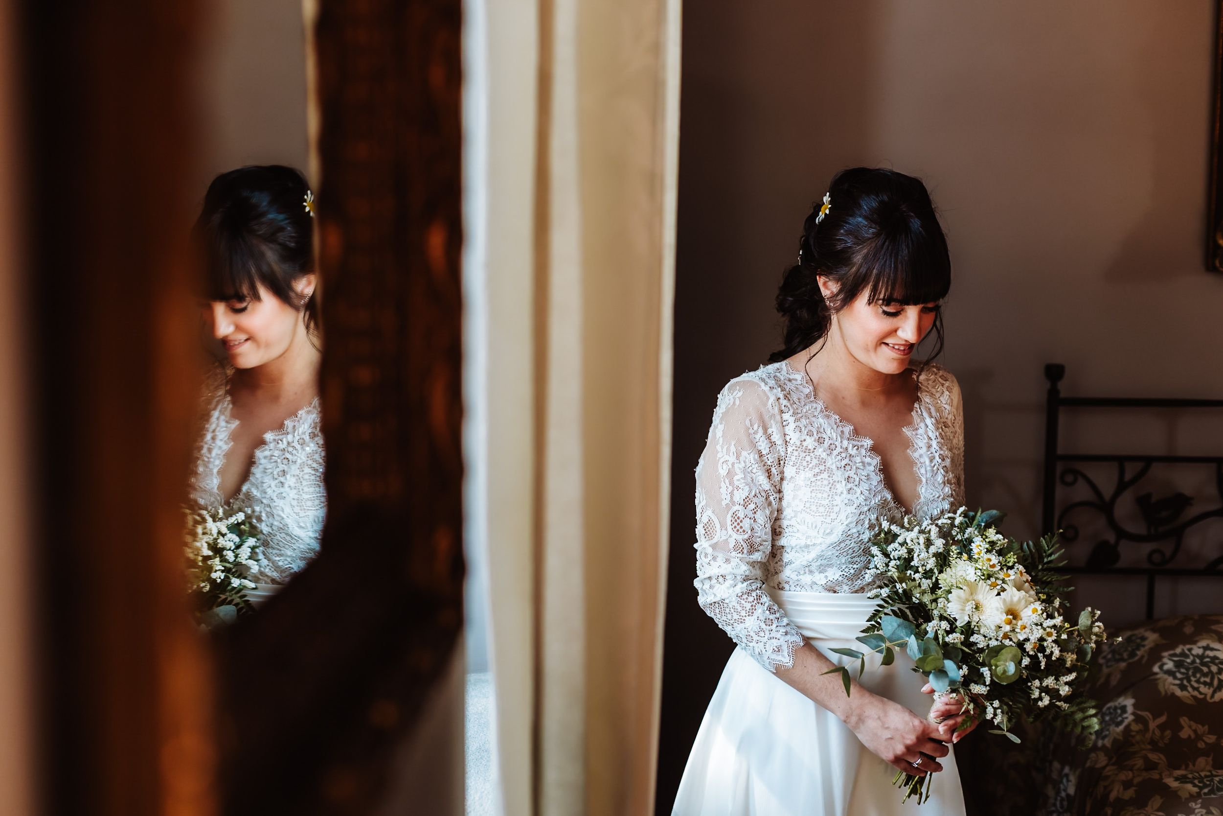 Bride in lace wedding gown holds white bouquet, reflected in mirror, soft natural light, elegant bridal portrait.