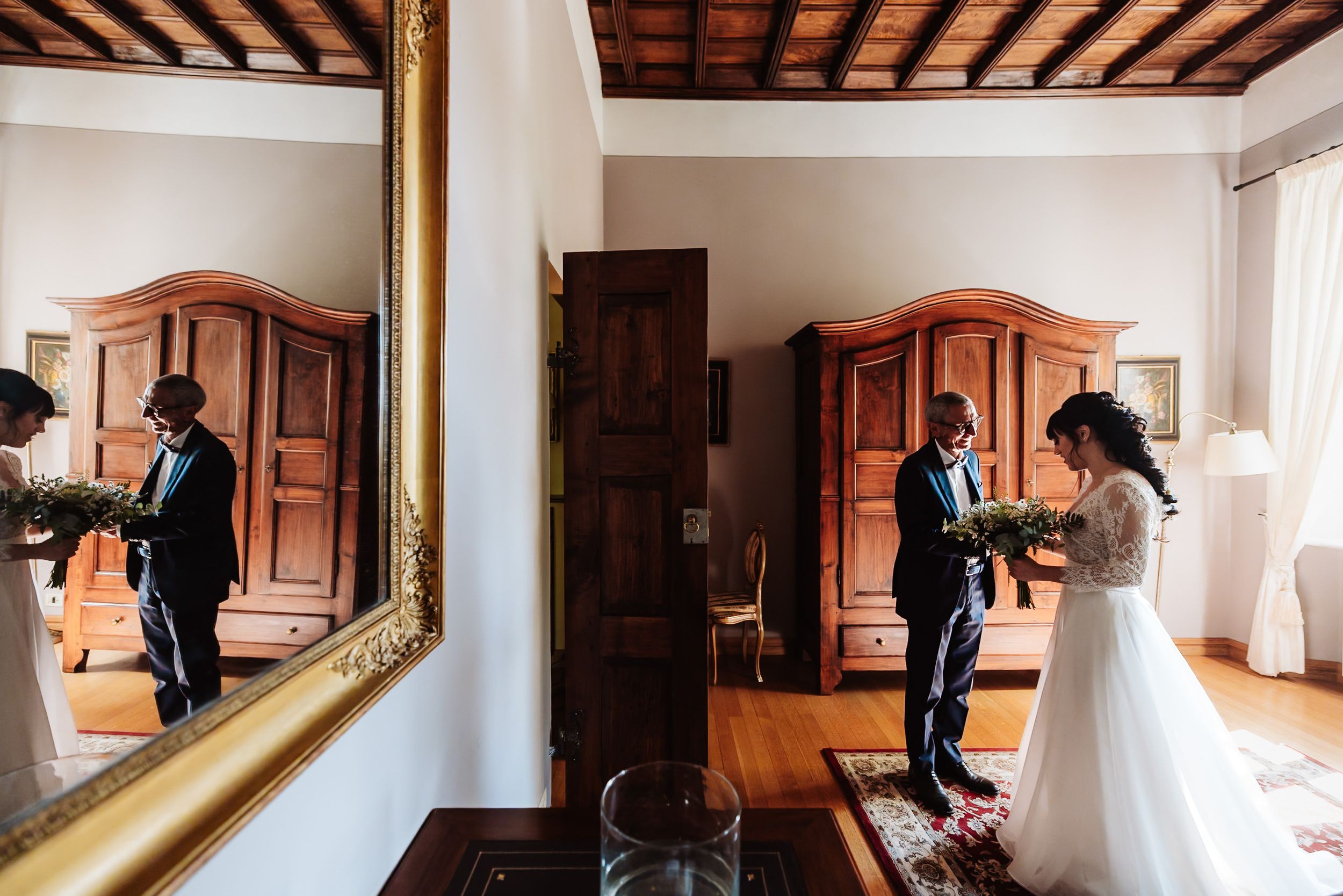 Bride and groom share a first look moment in an elegant room, reflected in a large ornate mirror.
