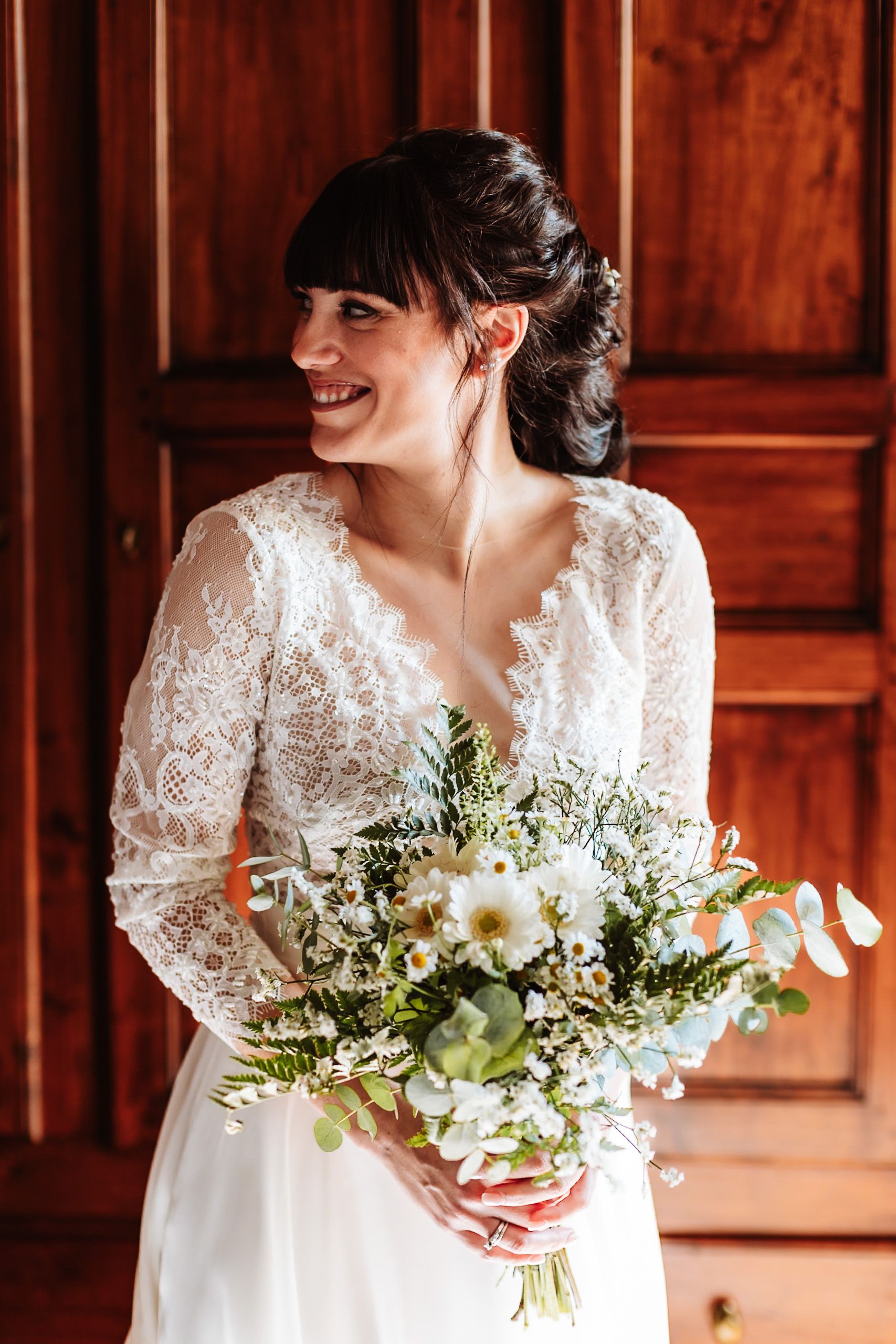 Smiling bride in lace long-sleeve wedding dress holding a white wildflower bouquet, standing before wooden doors.