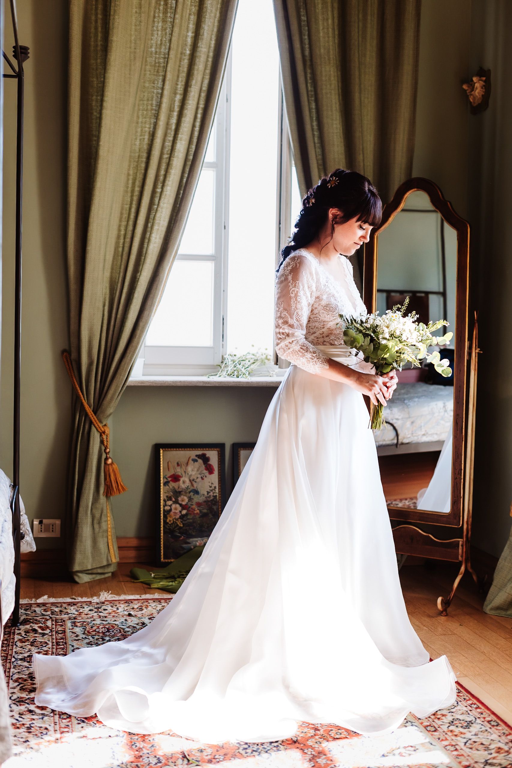Bride in white lace long-sleeve wedding gown holding bouquet by window with green curtains and vintage mirror.