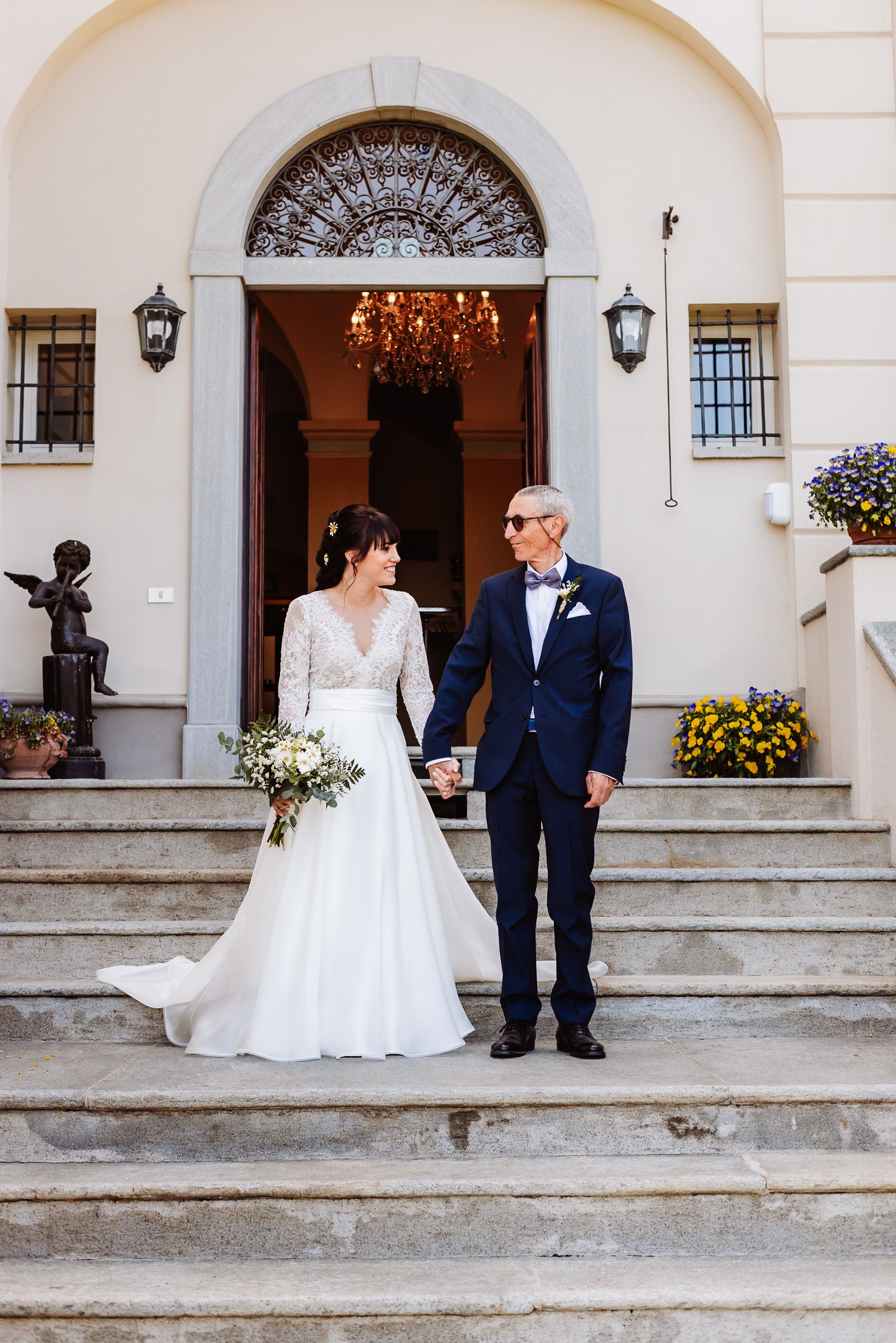 Bride and groom holding hands on stone steps outside elegant villa with arched doorway and chandelier.
