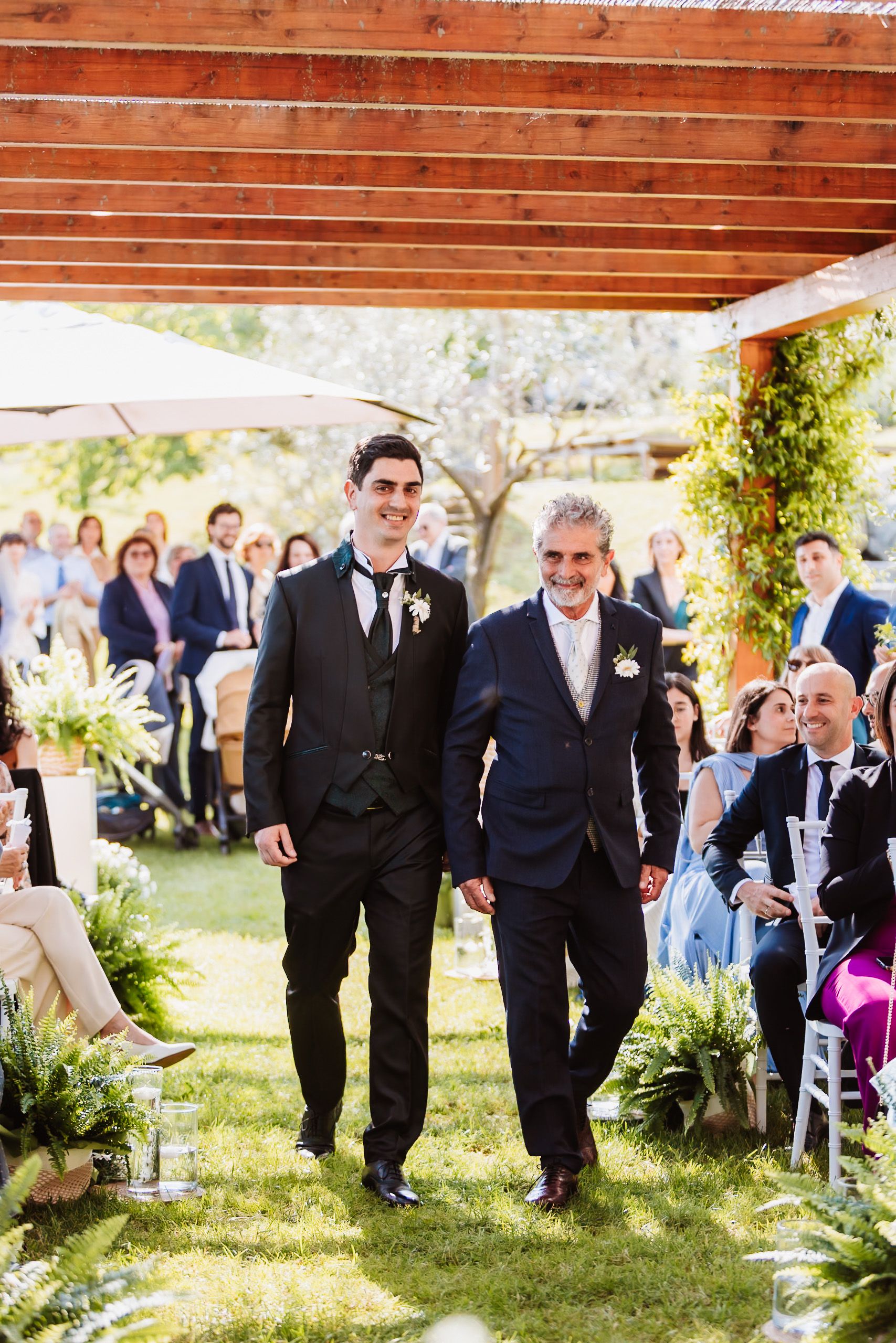 Two men in suits walk down the aisle at an outdoor wedding ceremony surrounded by guests and greenery.