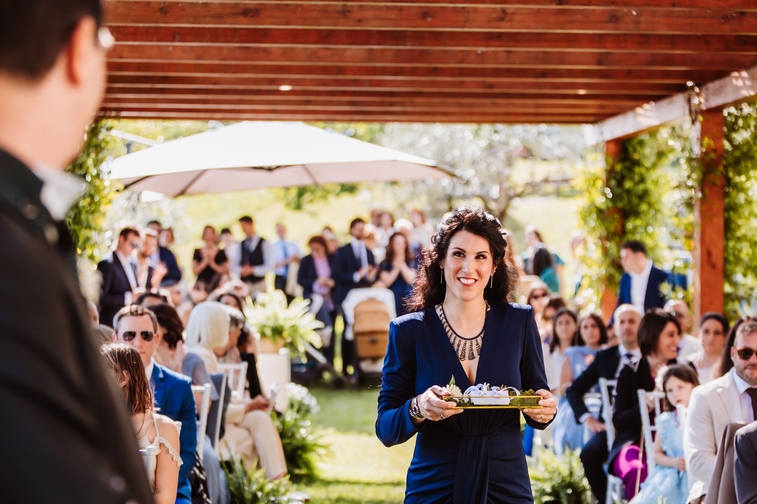 Woman in navy suit smiling while carrying a tray at an outdoor wedding reception with guests seated in background.