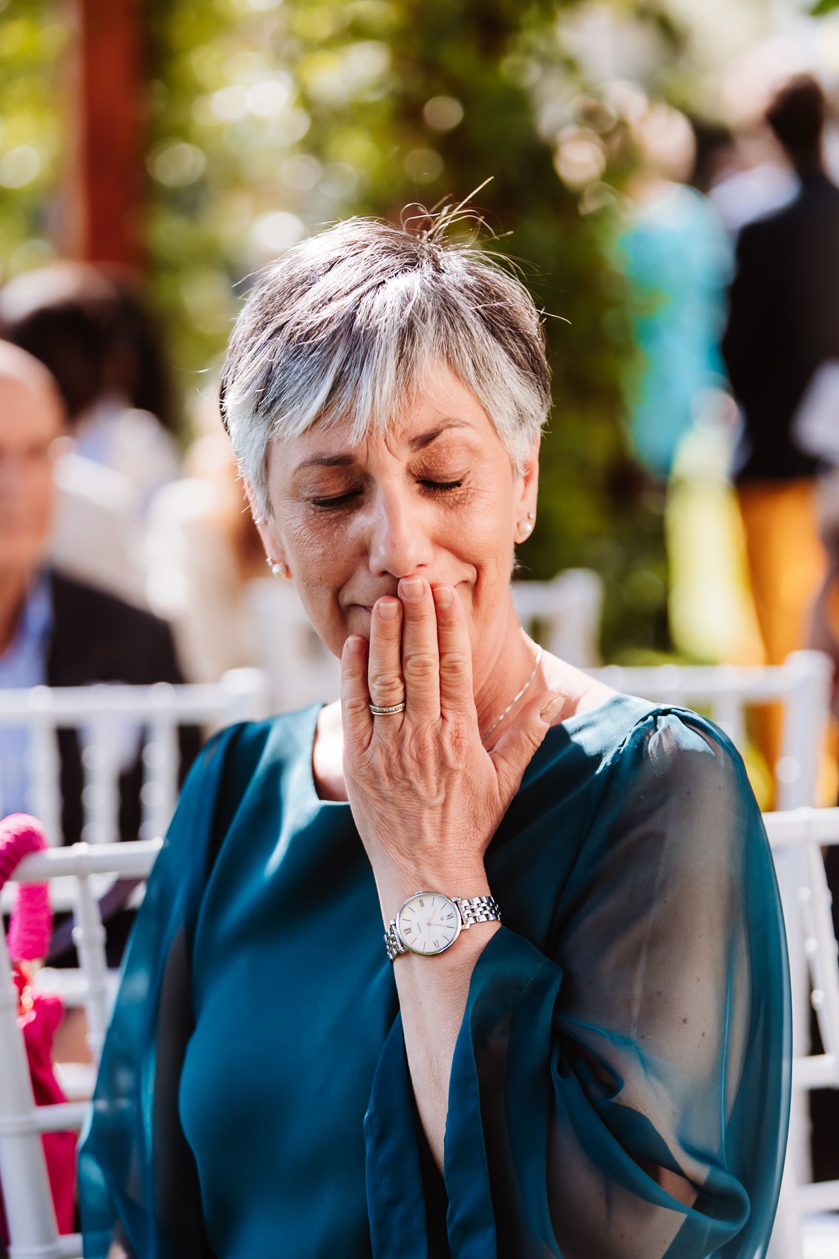 Emotional older woman with silver hair covering her mouth with her hand during an outdoor wedding ceremony.