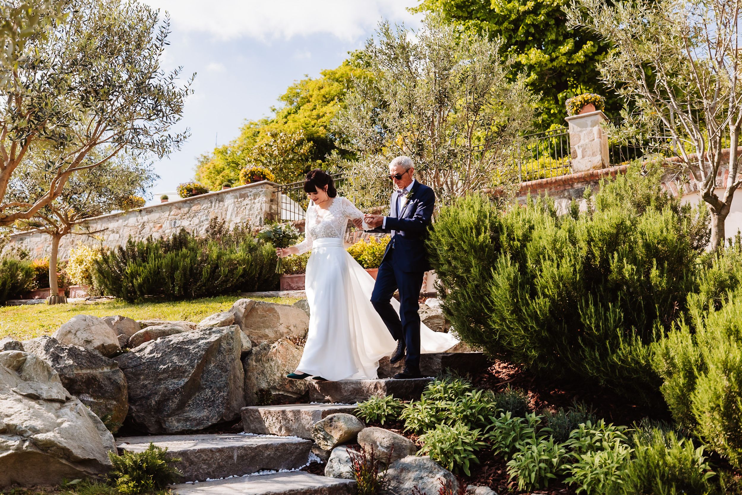 Bride and groom walking hand in hand through a lush garden with stone steps and greenery on their wedding day.