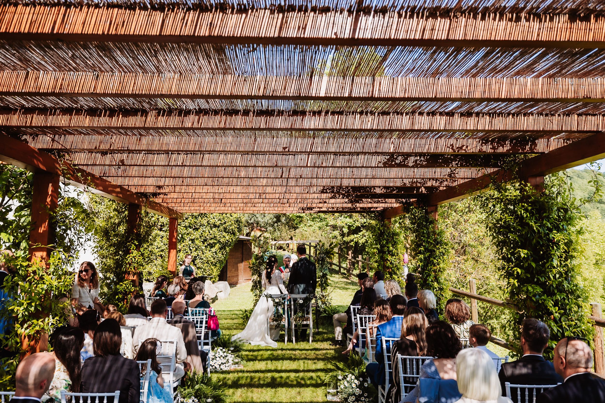 Outdoor wedding ceremony under a rustic pergola with reed roof, surrounded by lush greenery and seated guests.