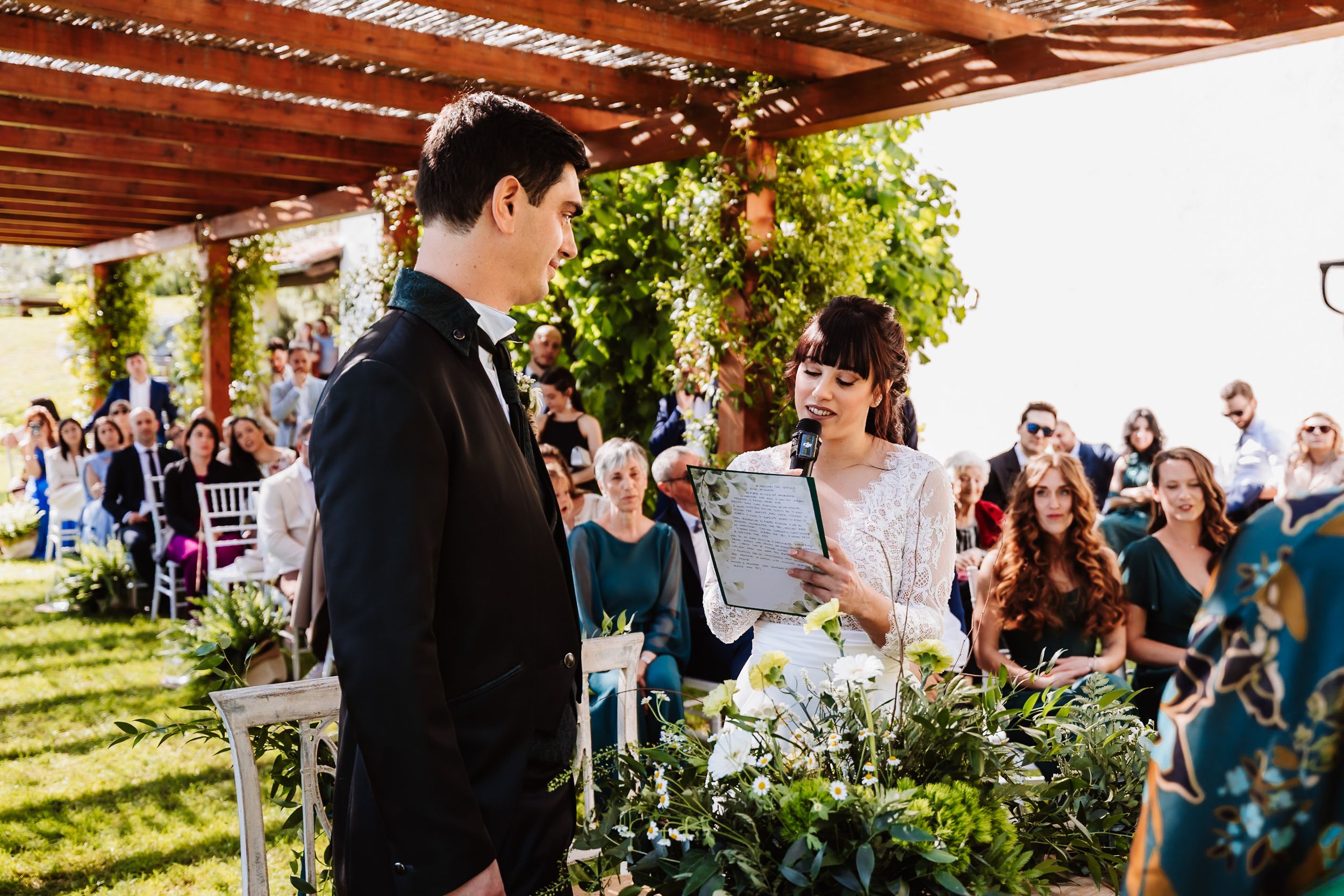 Bride reads vows to groom during outdoor wedding ceremony with guests seated in background.