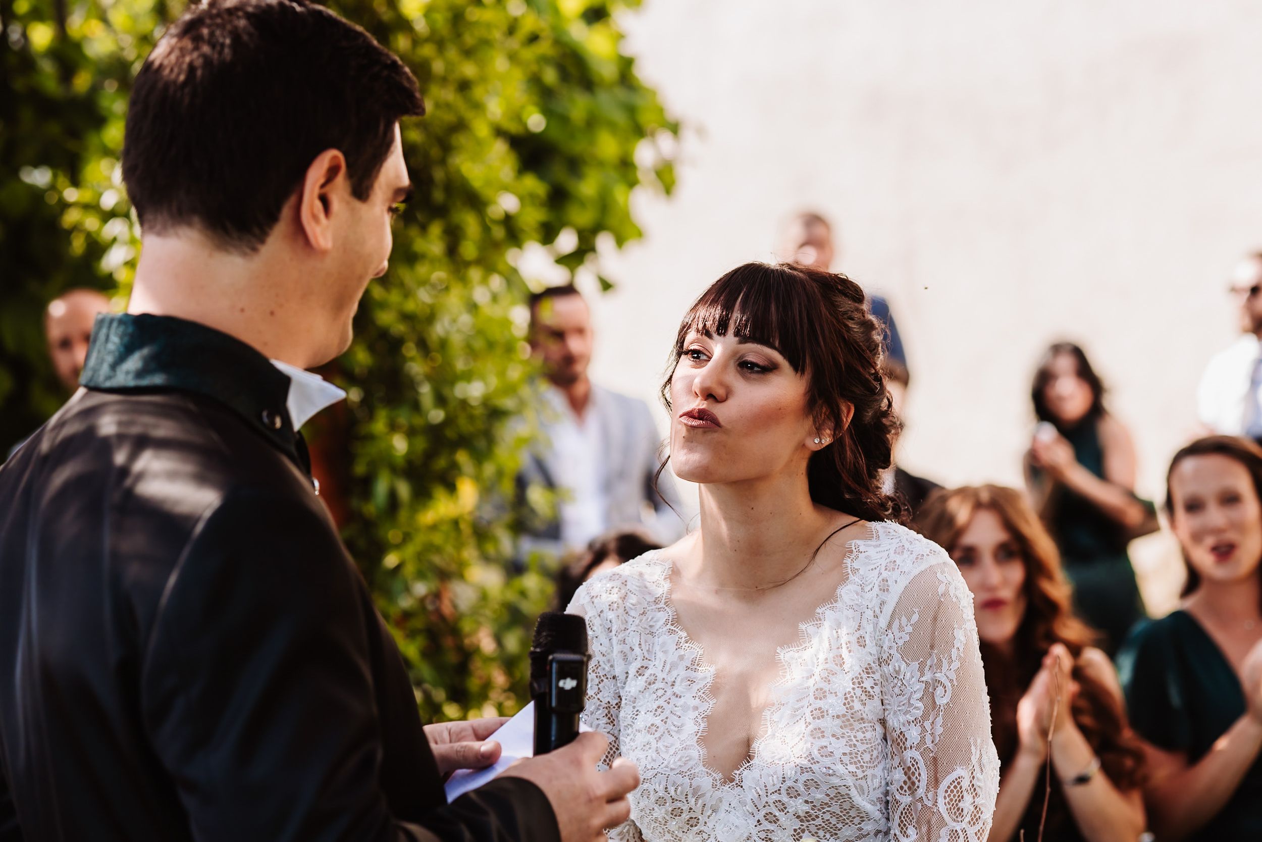 Bride in lace wedding dress exchanging vows with groom at outdoor wedding ceremony with guests in background.
