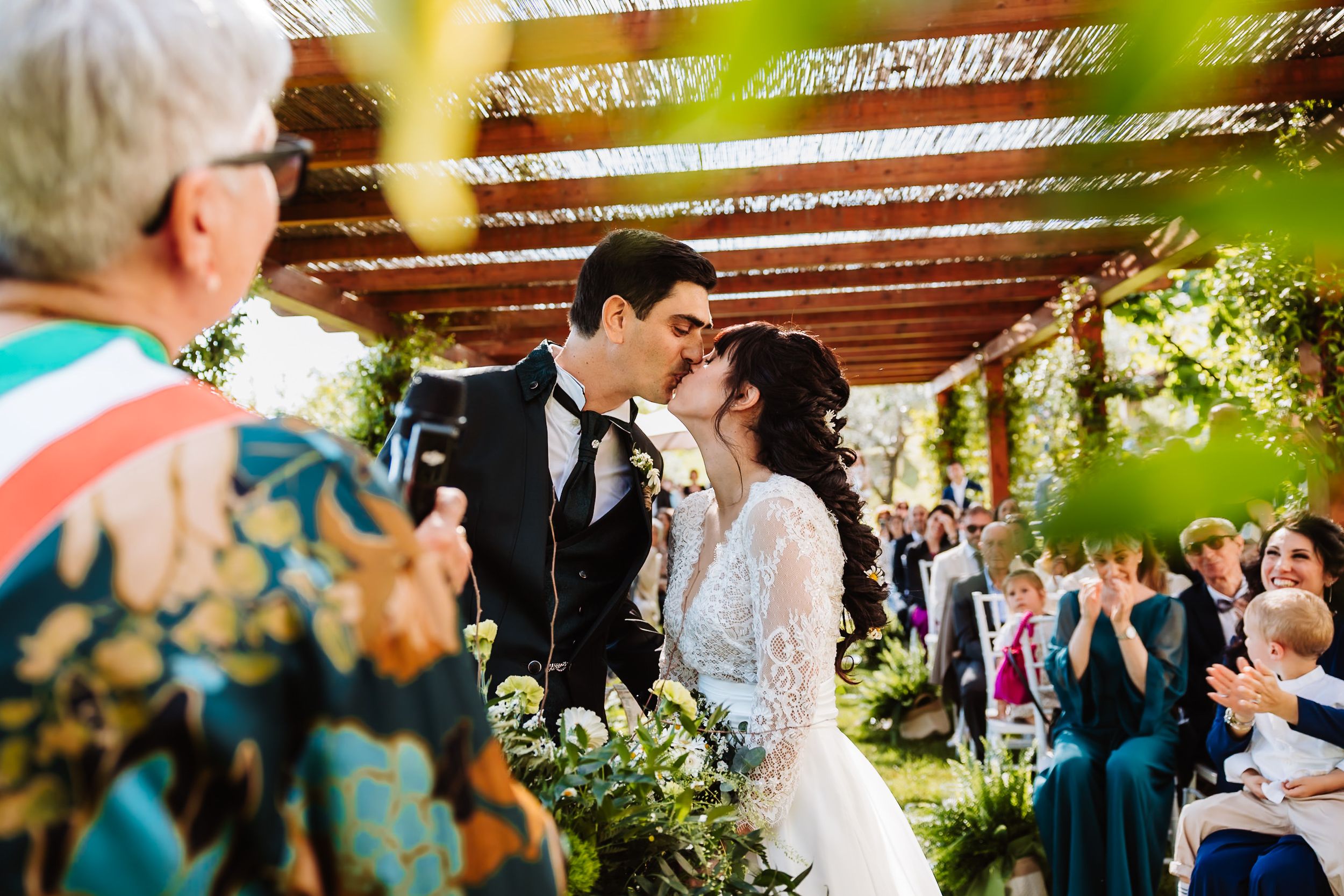 Bride and groom share first kiss at outdoor wedding ceremony under pergola with guests applauding.