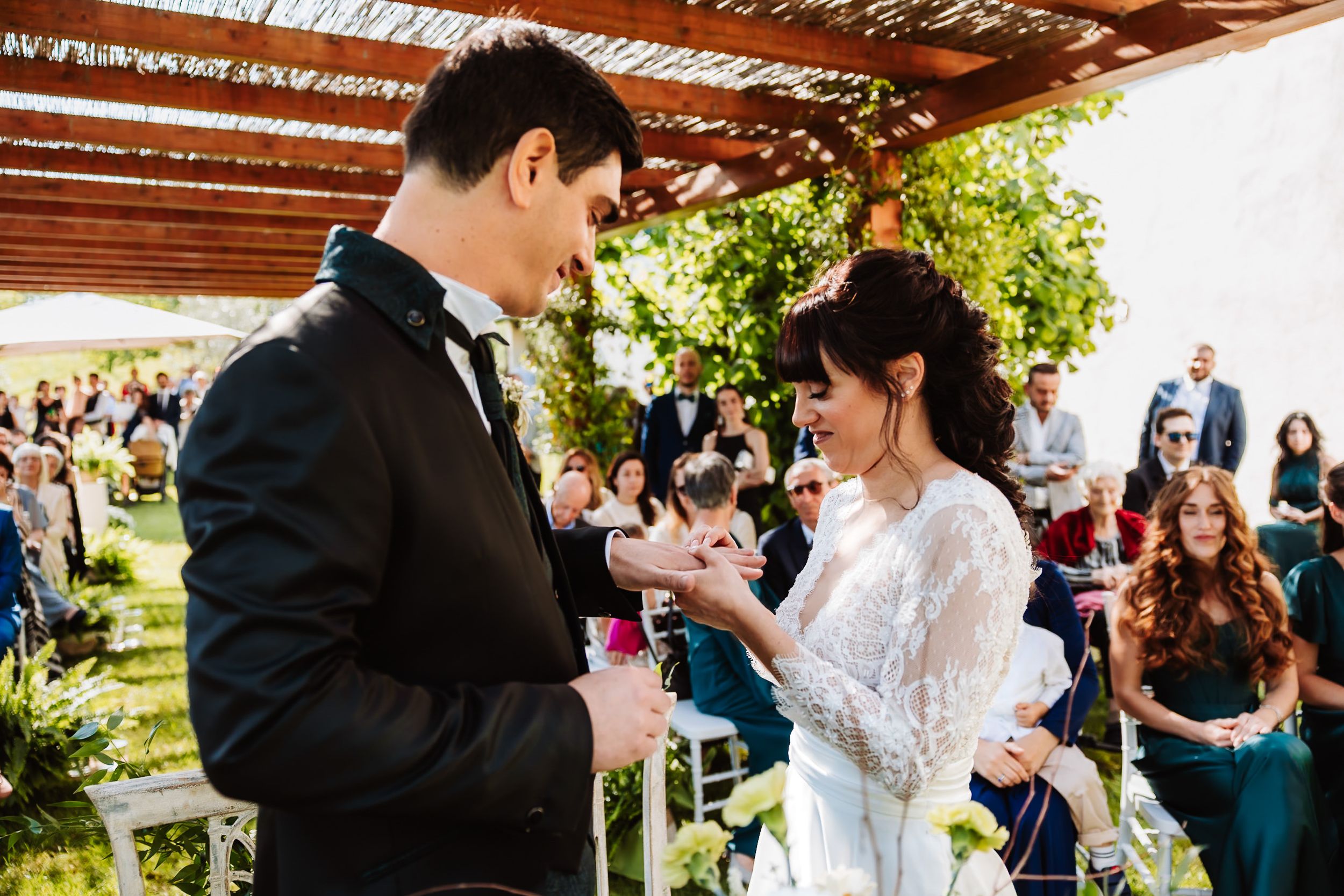 Bride and groom exchange rings during romantic outdoor wedding ceremony with smiling guests.