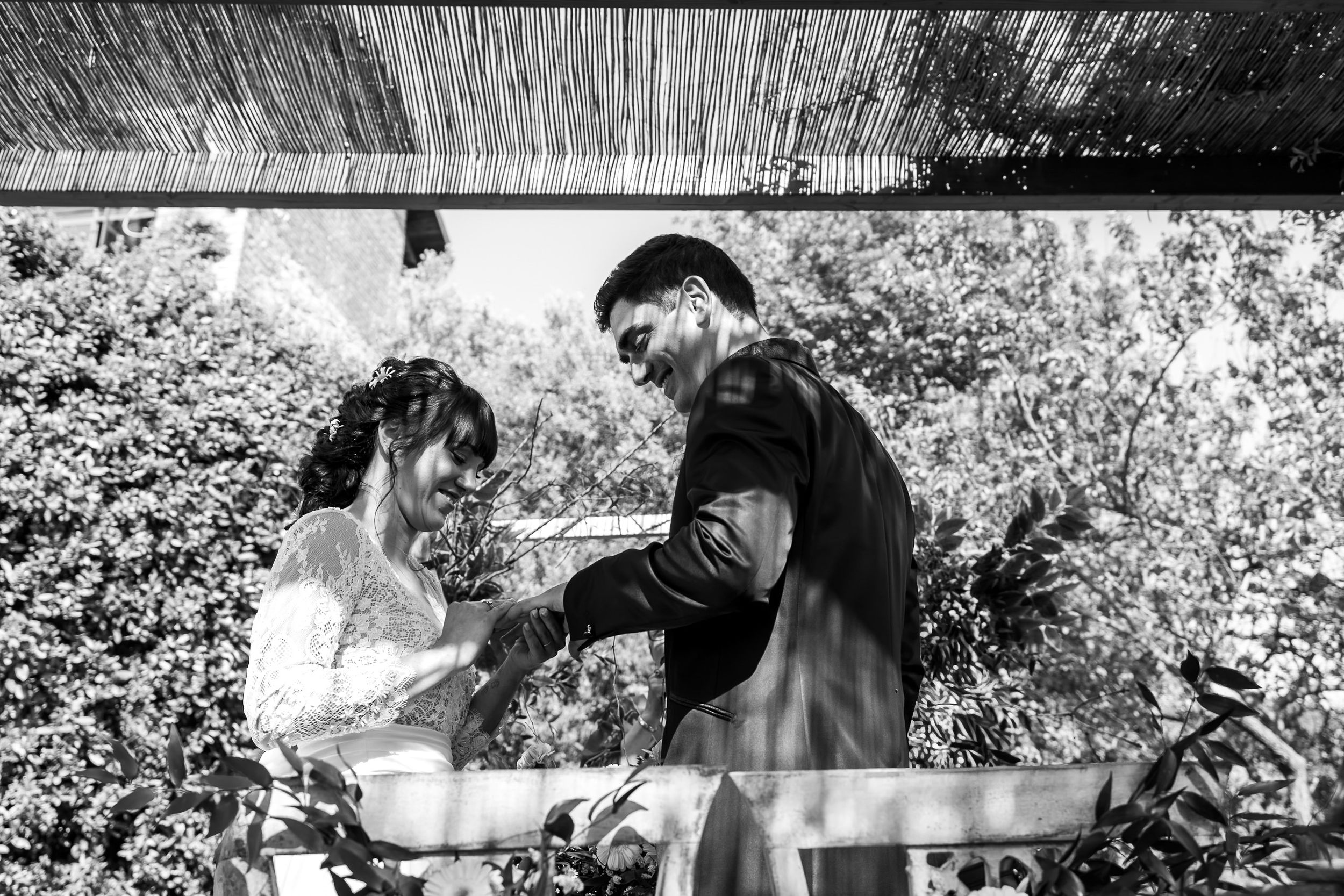 Black and white photo of a couple cutting their wedding cake outdoors under a pergola surrounded by lush greenery.