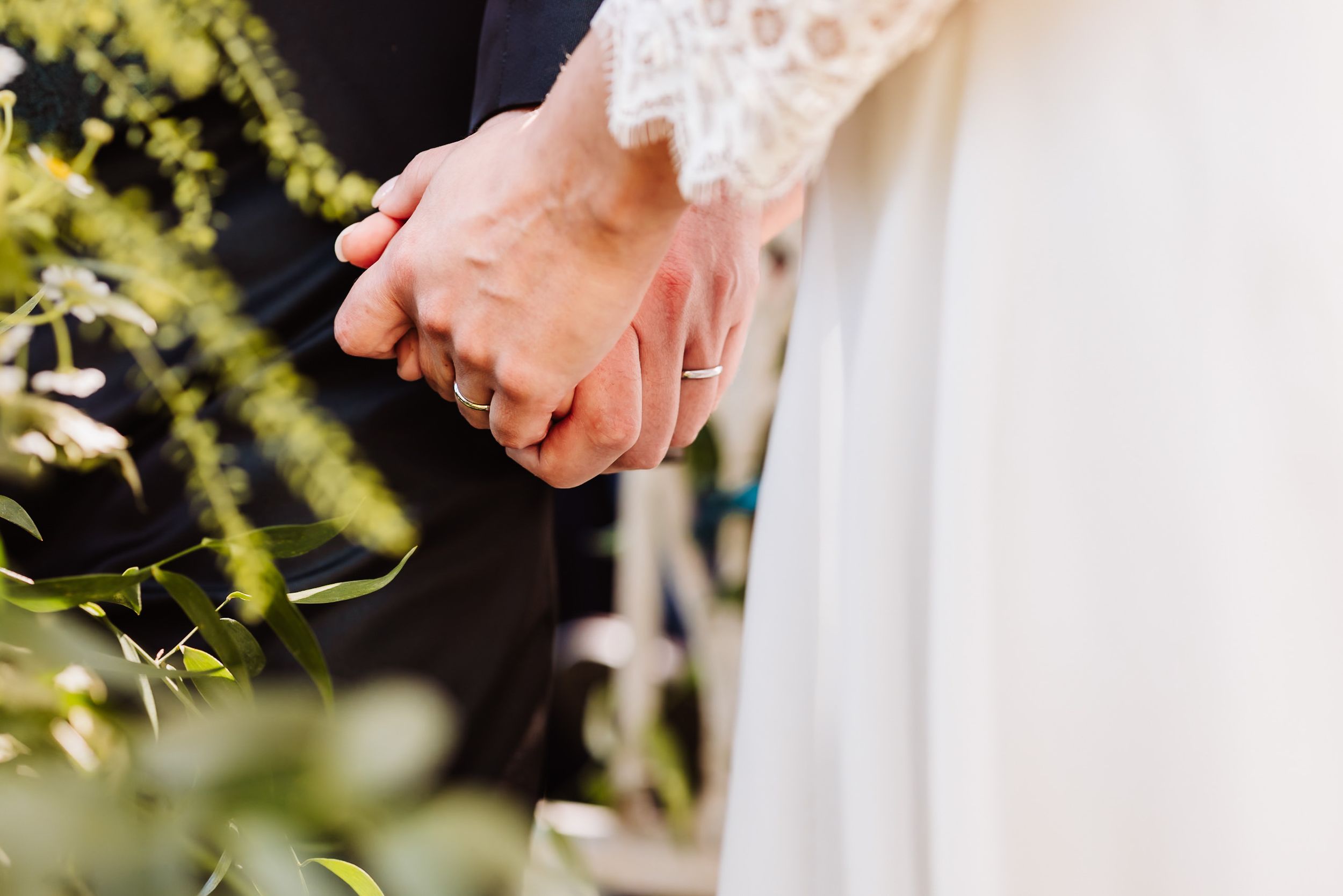 Close-up of bride and groom holding hands with wedding rings during outdoor ceremony.
