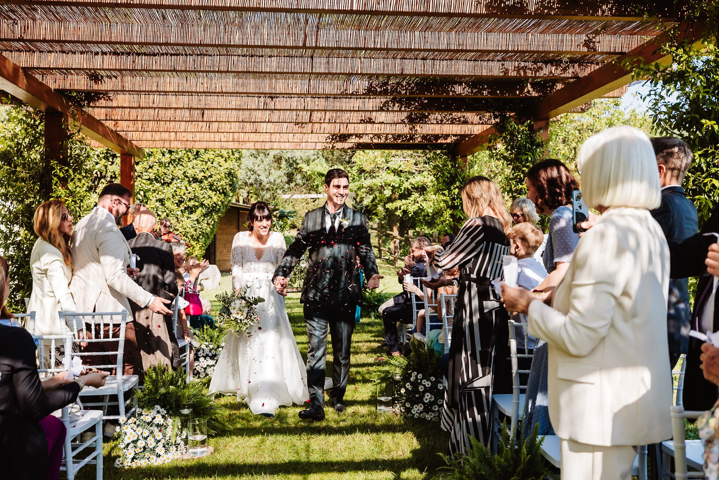 Bride and groom walk down outdoor wedding aisle under pergola surrounded by guests and lush greenery.