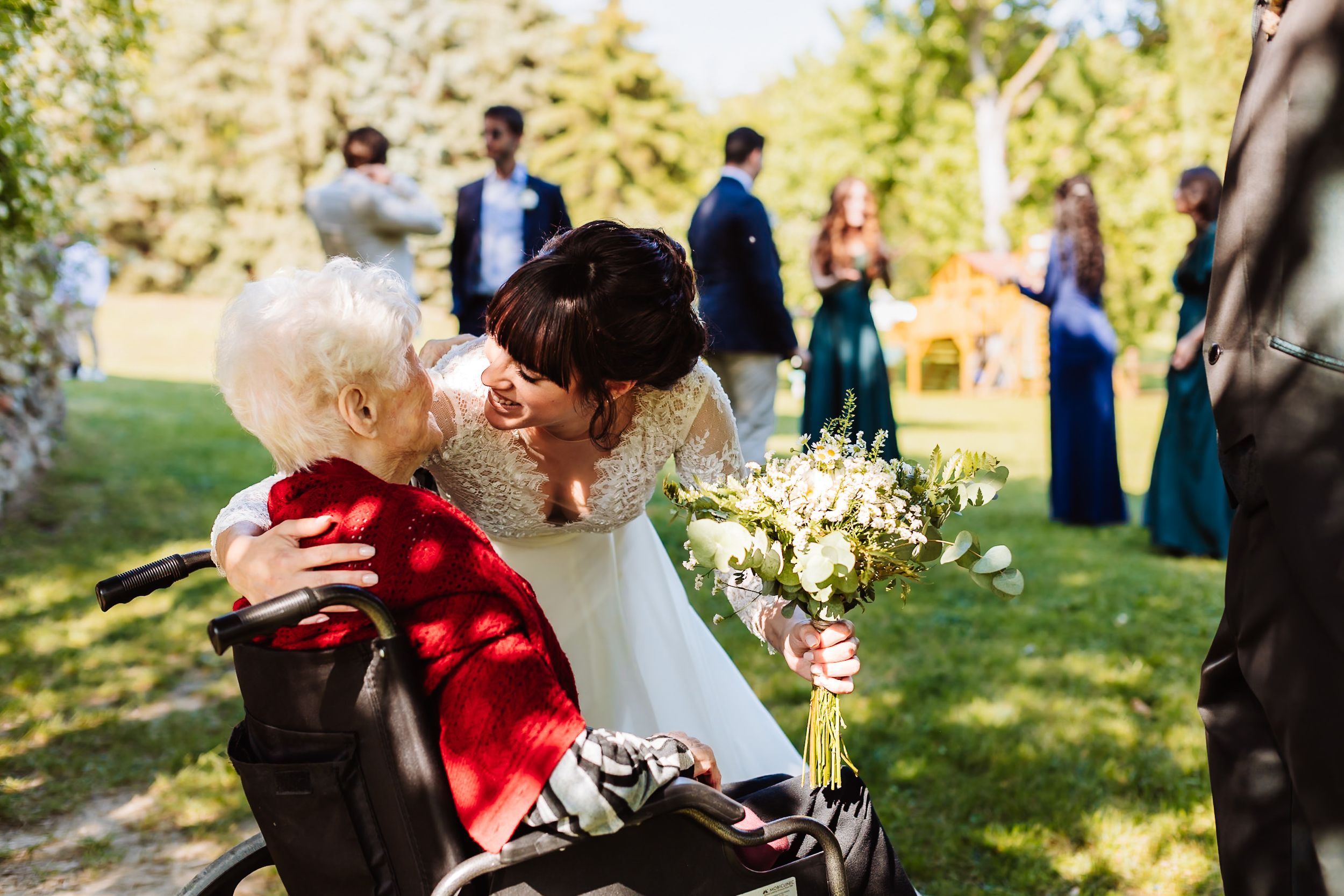 Bride in white gown kisses elderly woman in wheelchair at outdoor wedding ceremony.