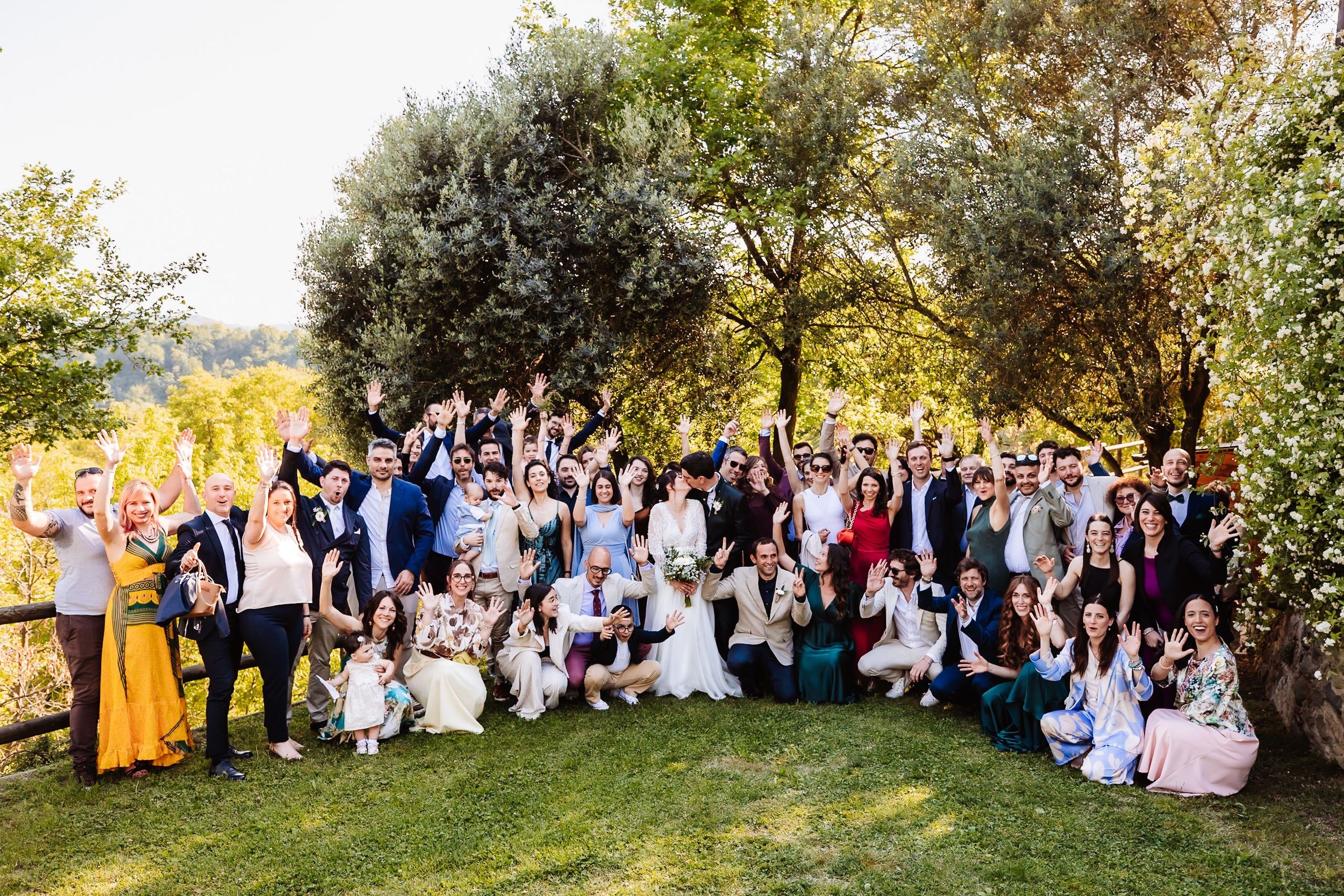 Large wedding group photo outdoors with bride and groom surrounded by smiling guests cheering under olive trees.