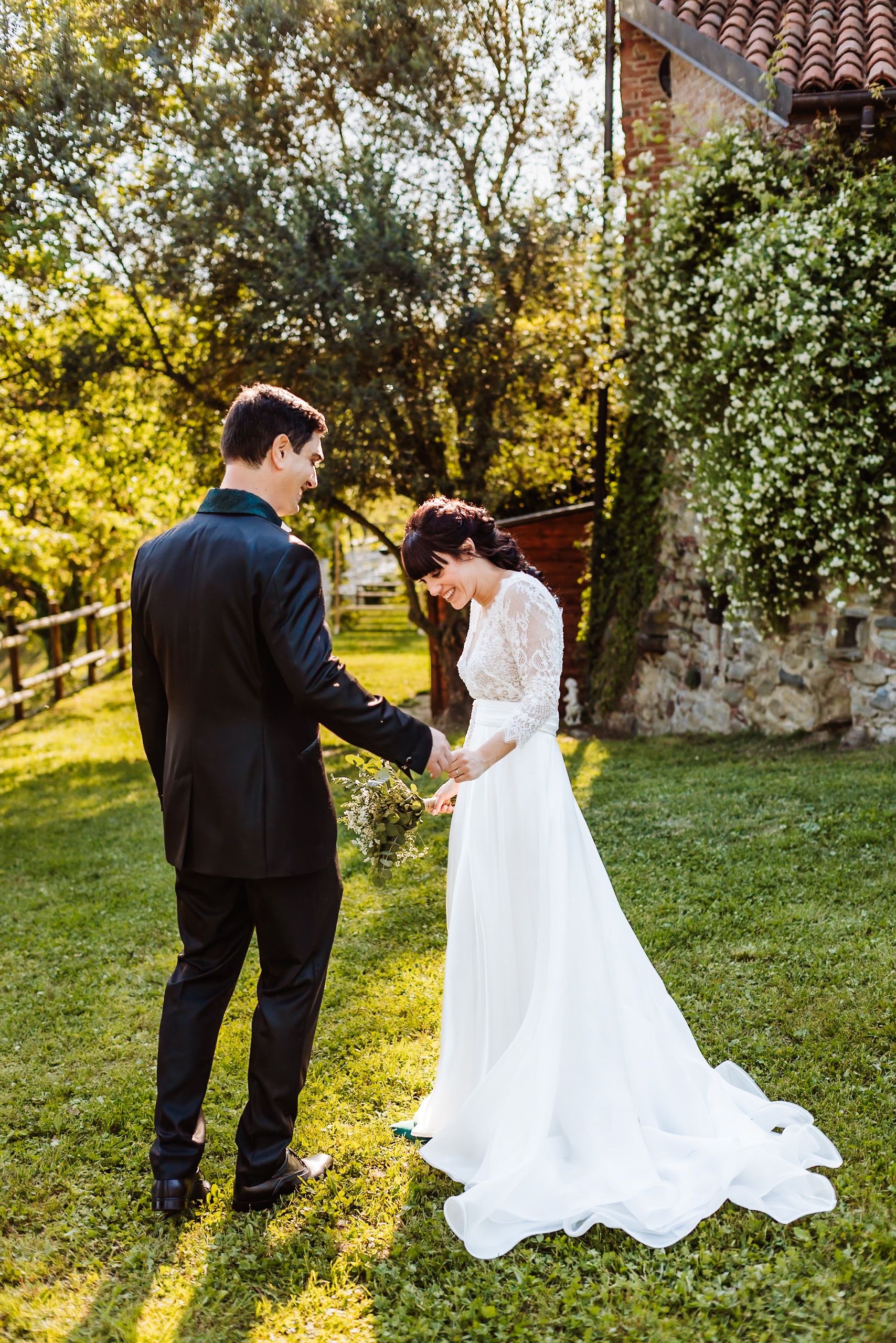Bride in white lace gown and groom in black suit hold hands in a sunlit garden near a rustic stone building.