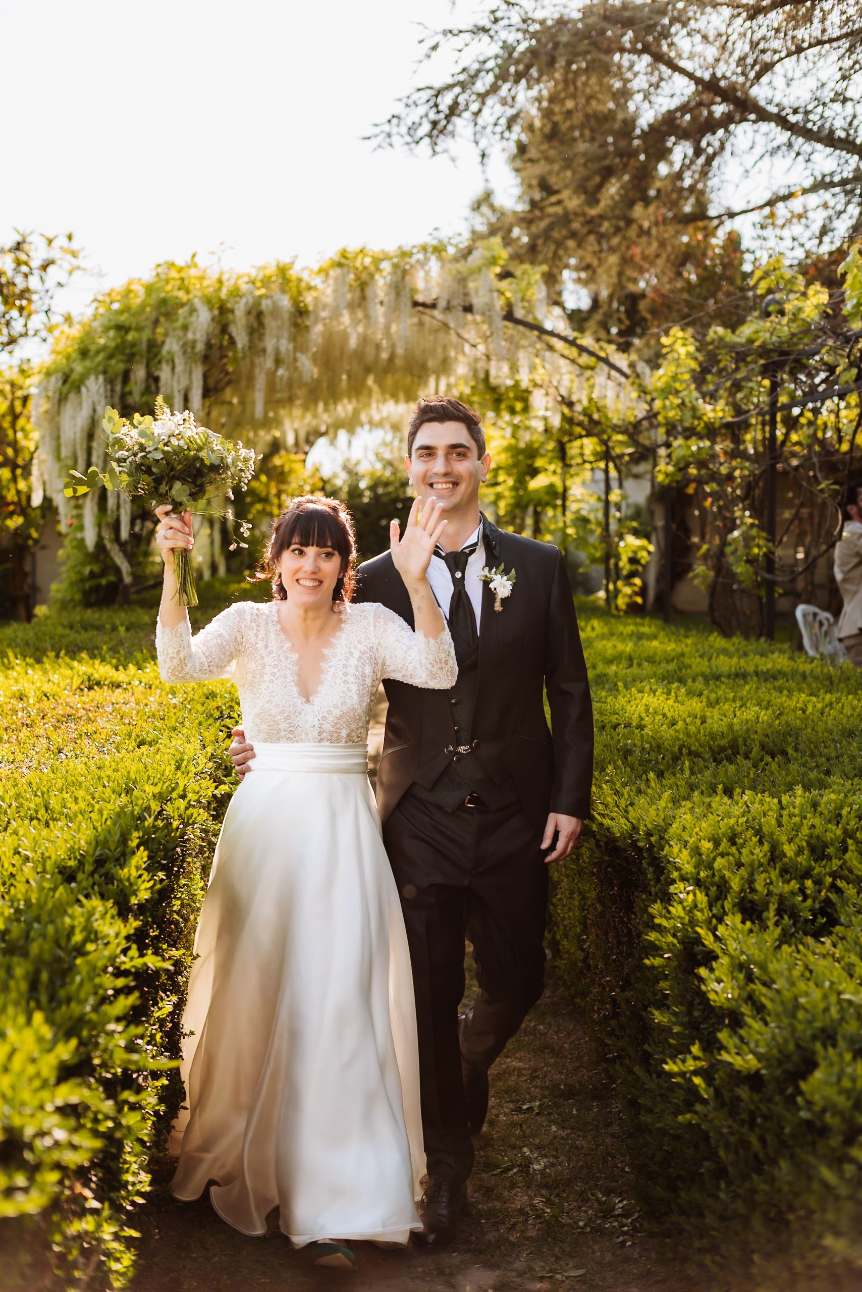Smiling bride and groom walking through garden hedge path, bride holding bouquet, groom waving, white wisteria overhead.