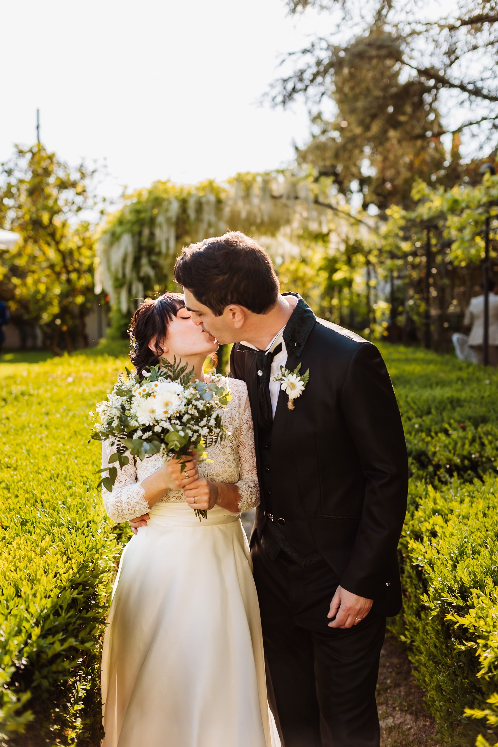A bride and groom share a romantic kiss in a sunlit garden, she holds a white bouquet while he wears a black tuxedo.