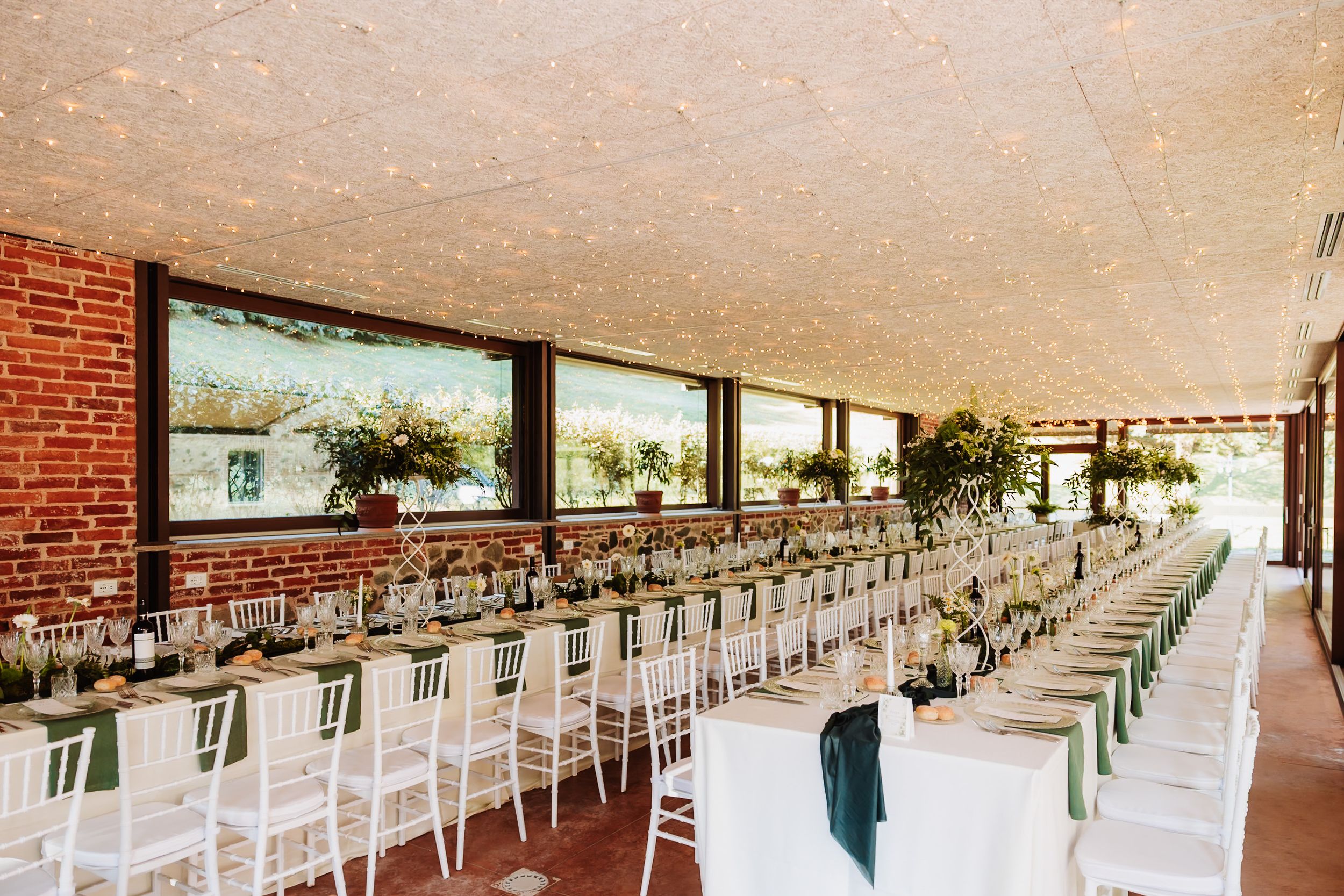 Elegant wedding reception hall with long white tables, green decor, and brick walls under a textured ceiling.