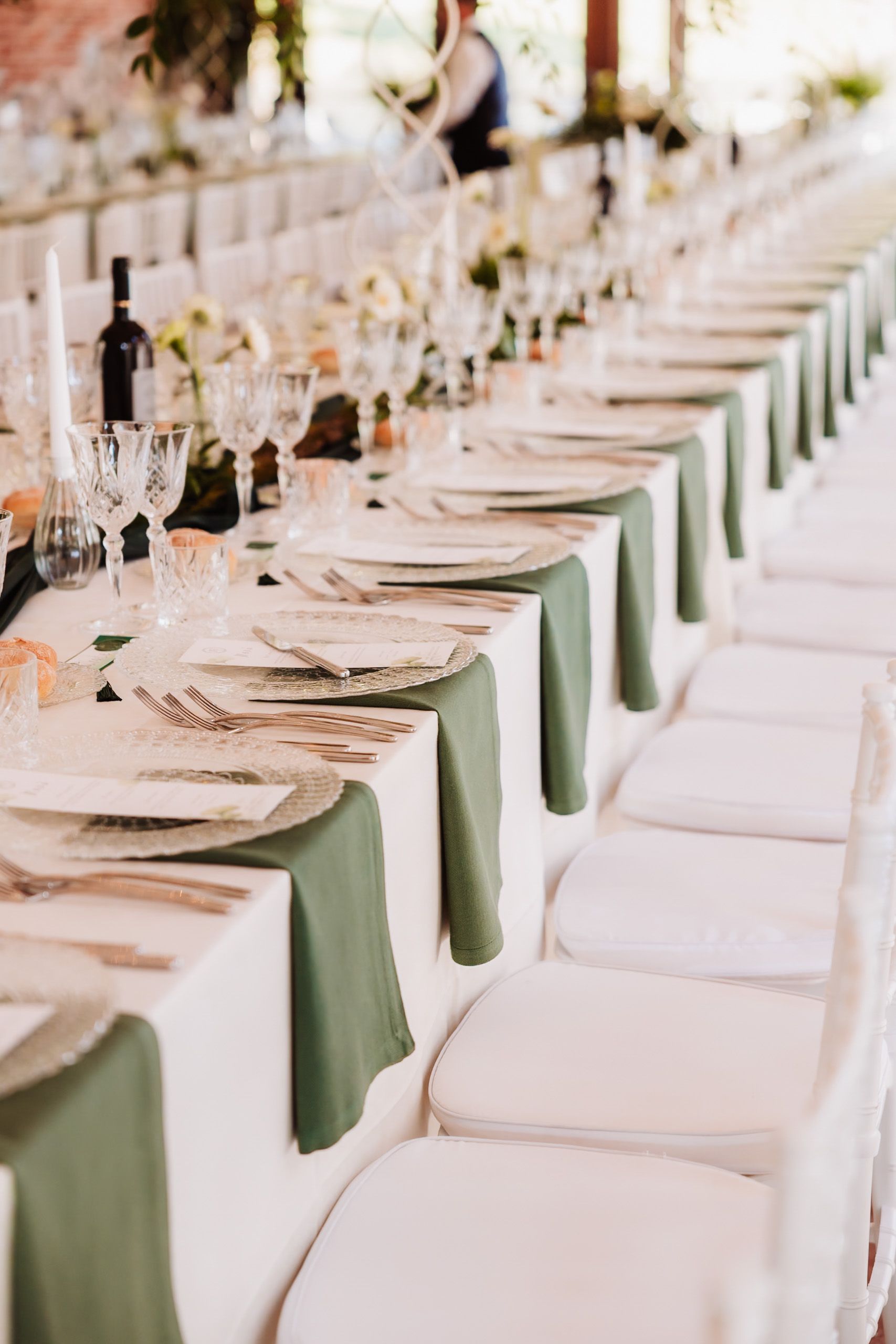 Elegant wedding reception table with green napkins, crystal glasses, and white chairs in a long formal dining arrangement.