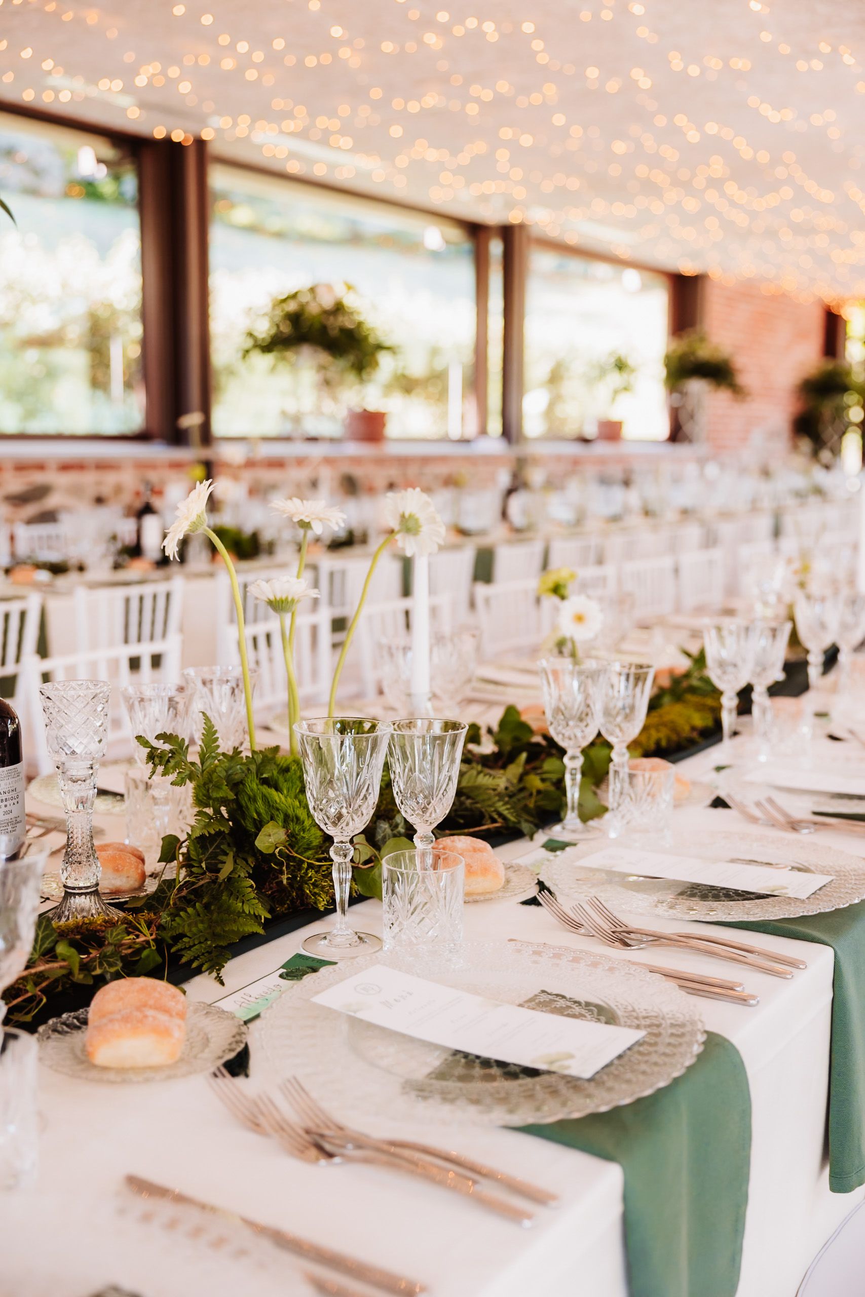 Elegant wedding reception table with crystal glassware, greenery runner, white flowers, and fairy lights overhead.