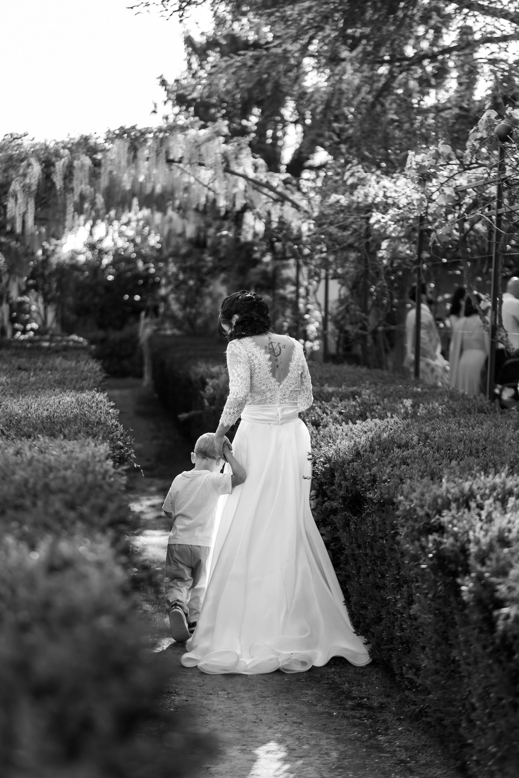 Bride in lace wedding gown walks hand-in-hand with child through garden path lined with manicured hedges, black and white photo.
