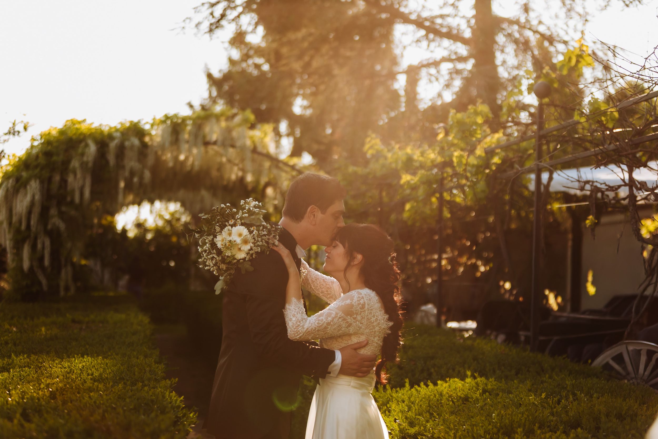 Couple sharing a romantic kiss at golden hour in a lush garden, bride holding white flowers in a elegant wedding dress.