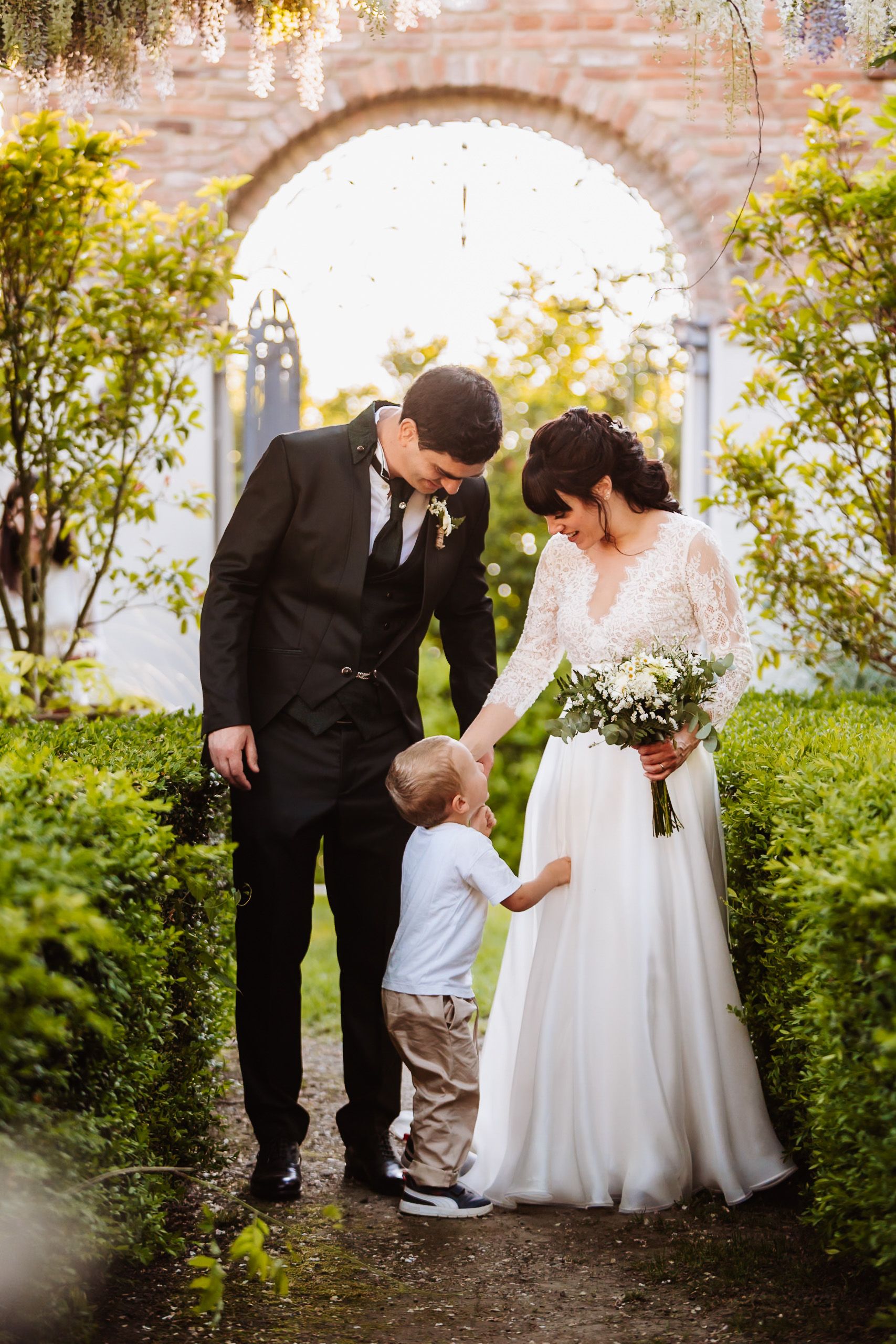 Bride and groom in garden arch holding hands with young child on wedding day.