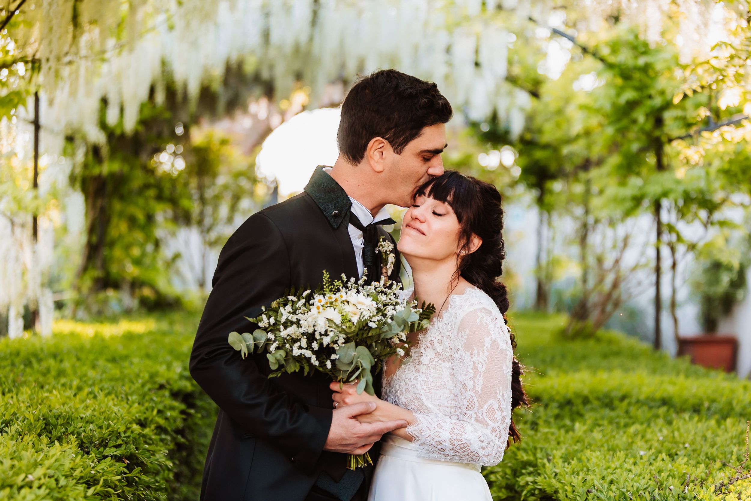 Bride and groom share a romantic kiss under white wisteria flowers in a lush green garden wedding setting.