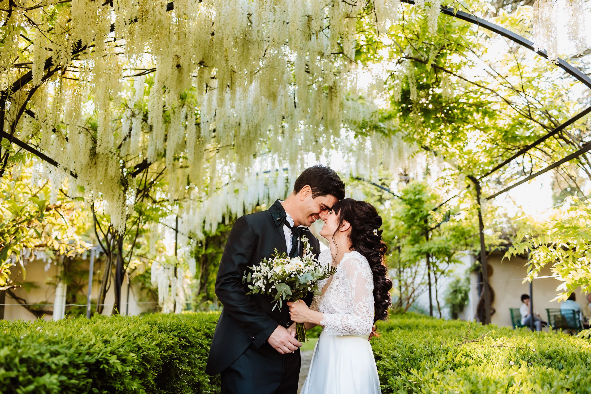Bride and groom share romantic moment under hanging white wisteria flowers in lush garden, groom holds bouquet.