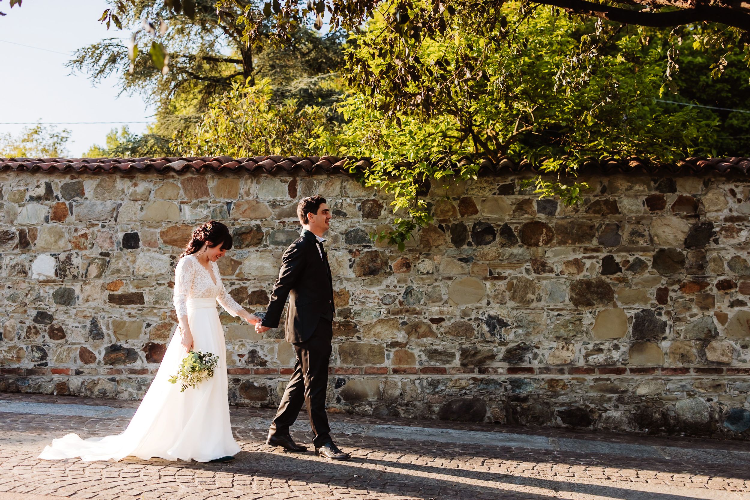 A bride and groom walk hand in hand along a rustic stone wall surrounded by lush green trees on their wedding day.