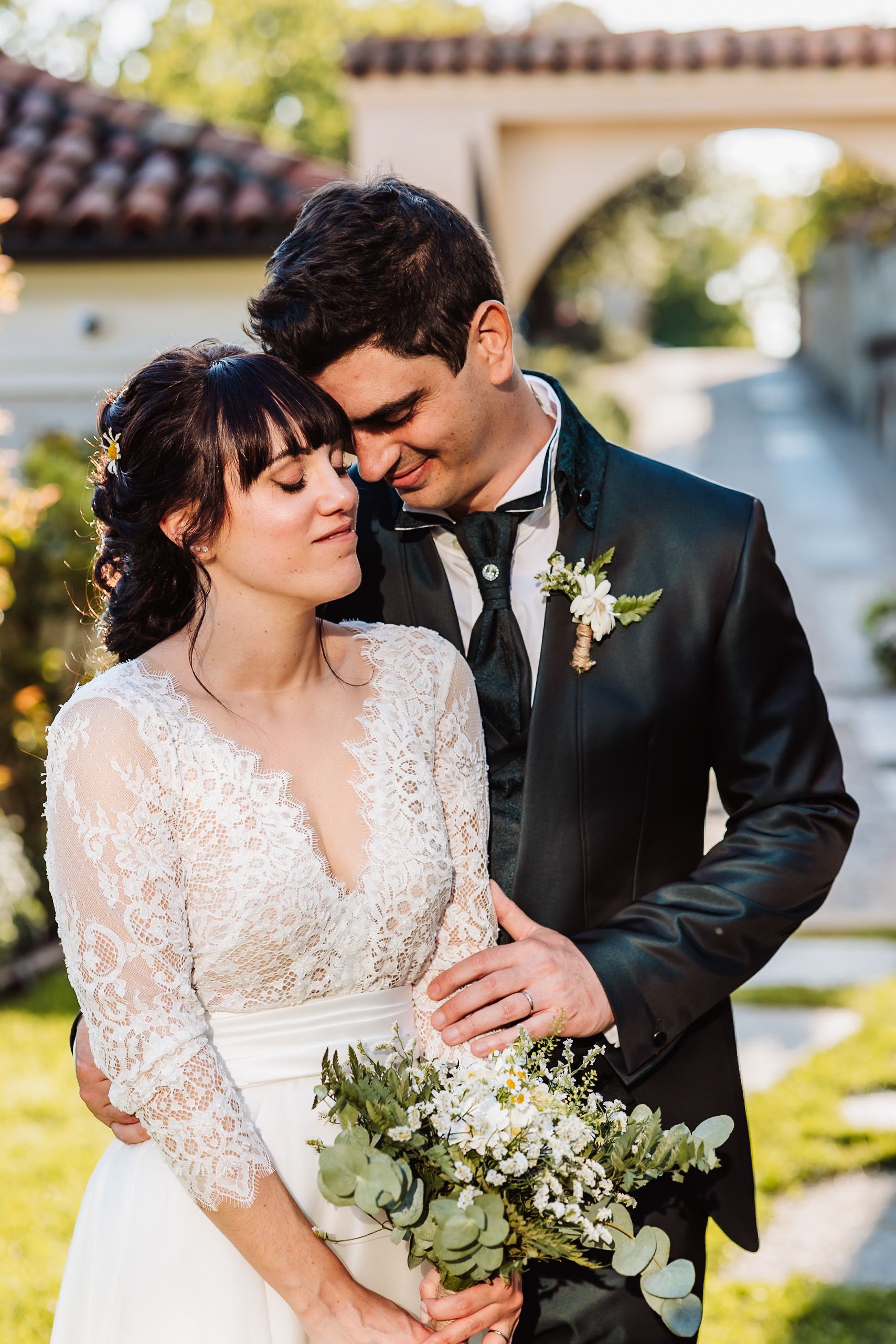Bride in lace gown holding greenery bouquet leans into groom in black tuxedo outdoors at wedding venue.