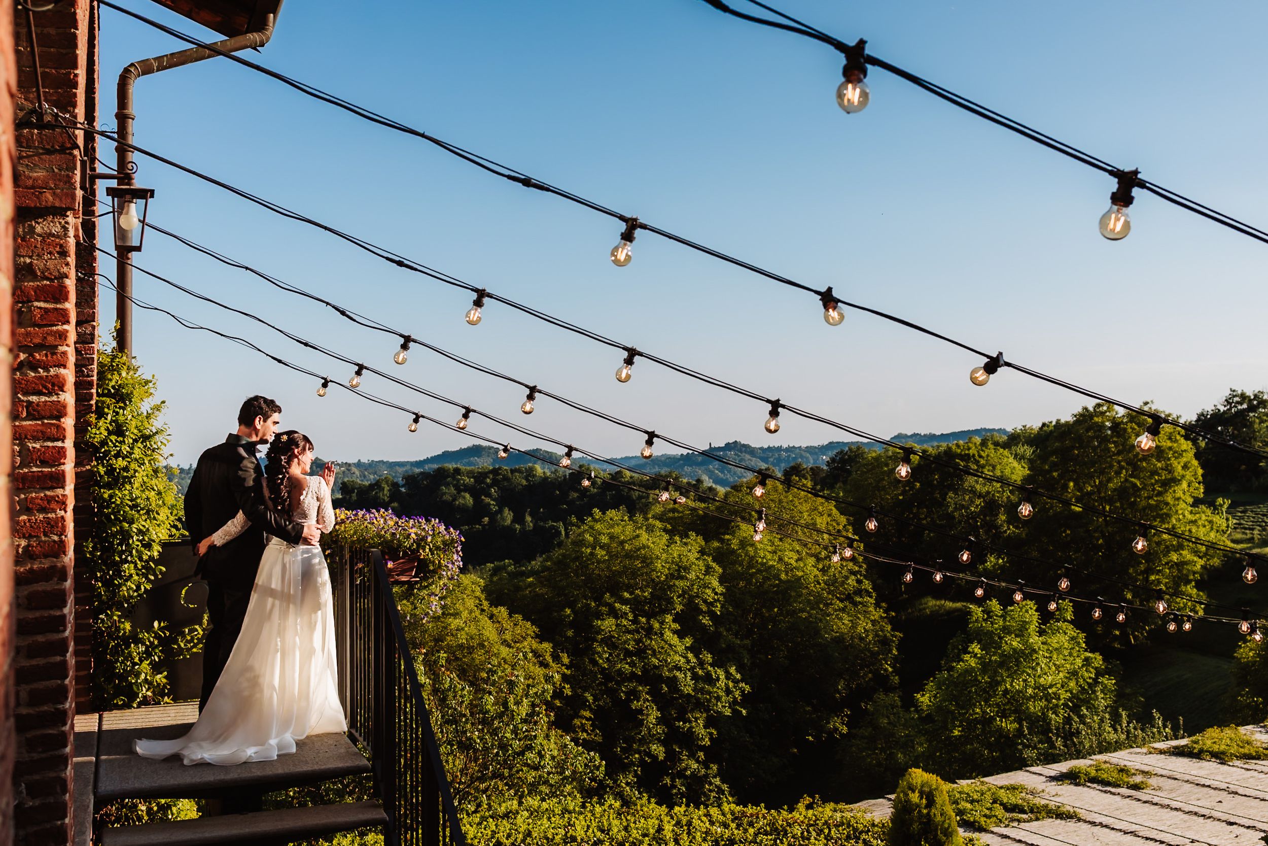 Bride and groom embrace on a rustic balcony with string lights, overlooking lush green hills at dusk.