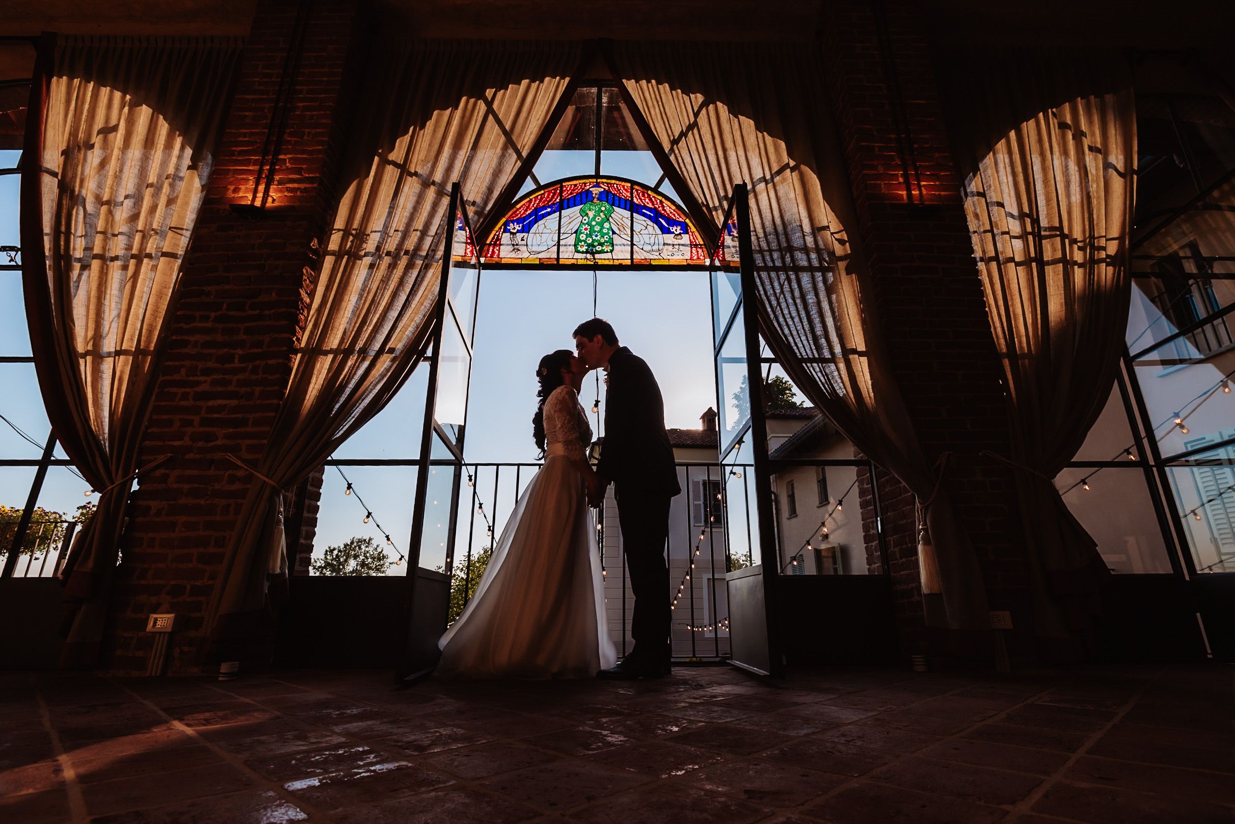 Silhouette of bride and groom sharing a kiss framed by stained glass window and draped curtains in elegant venue.