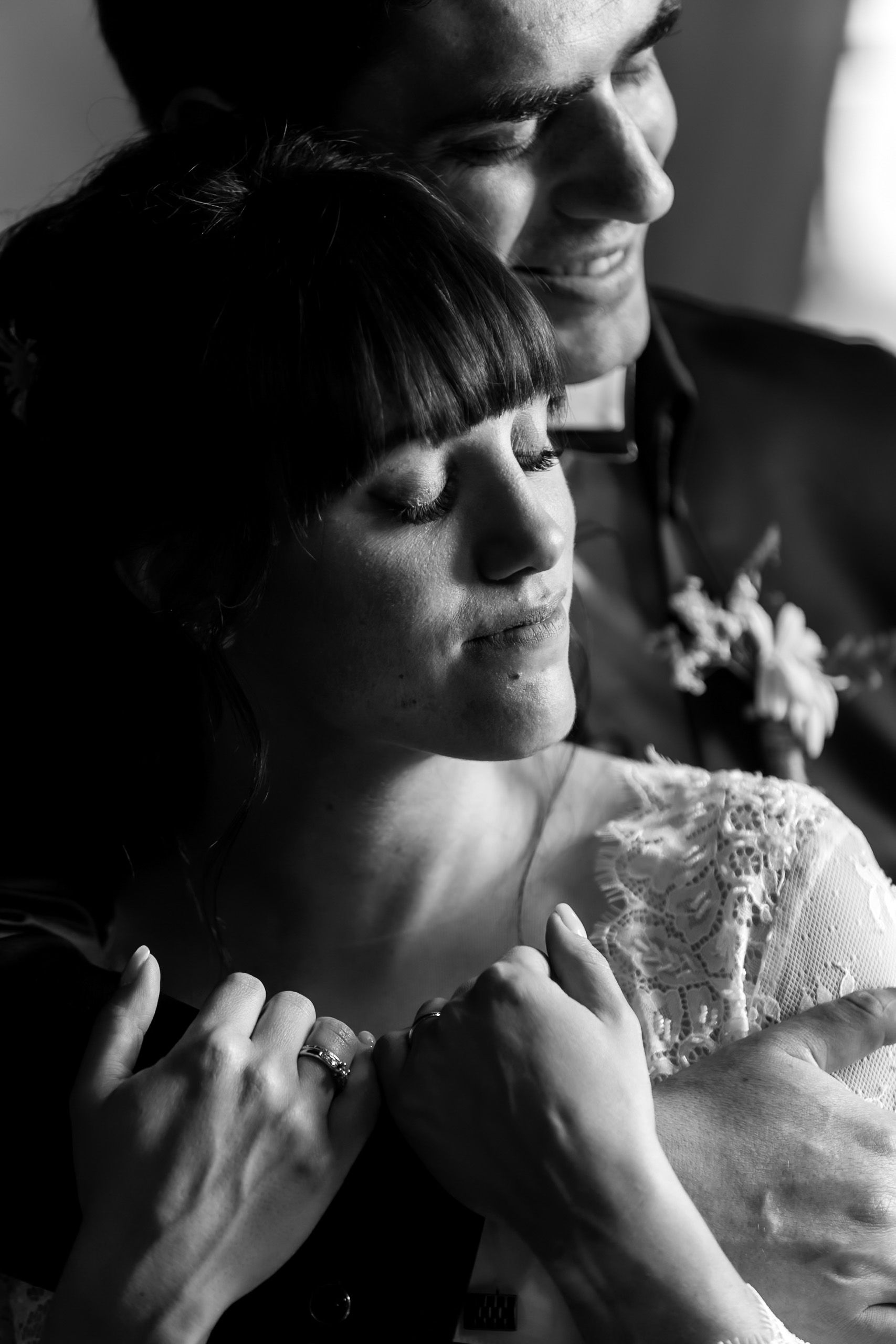 Black and white wedding photo of bride and groom embracing tenderly, bride wearing lace dress and ring.