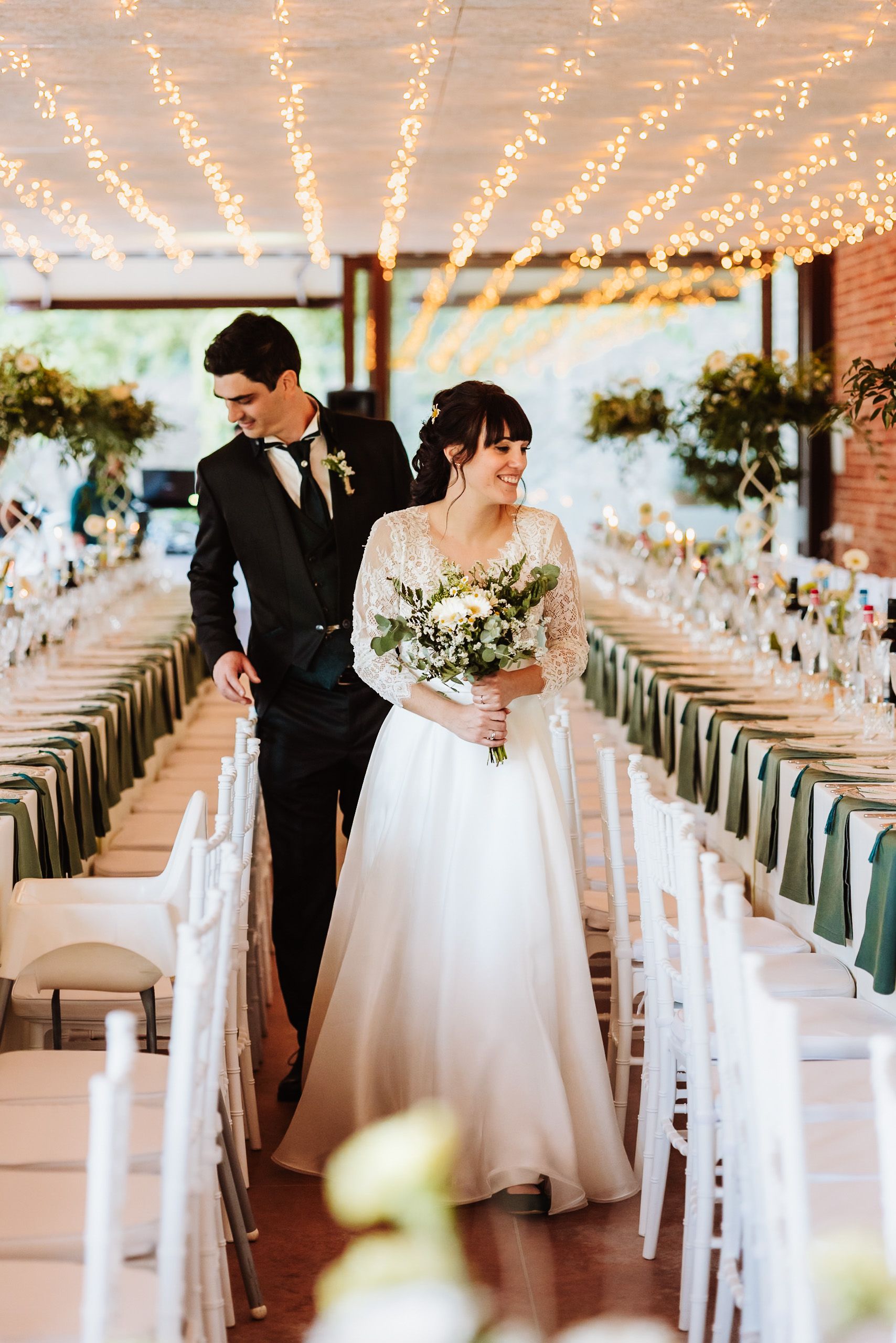 Bride and groom walk through elegantly decorated wedding reception hall with fairy lights and green table settings.