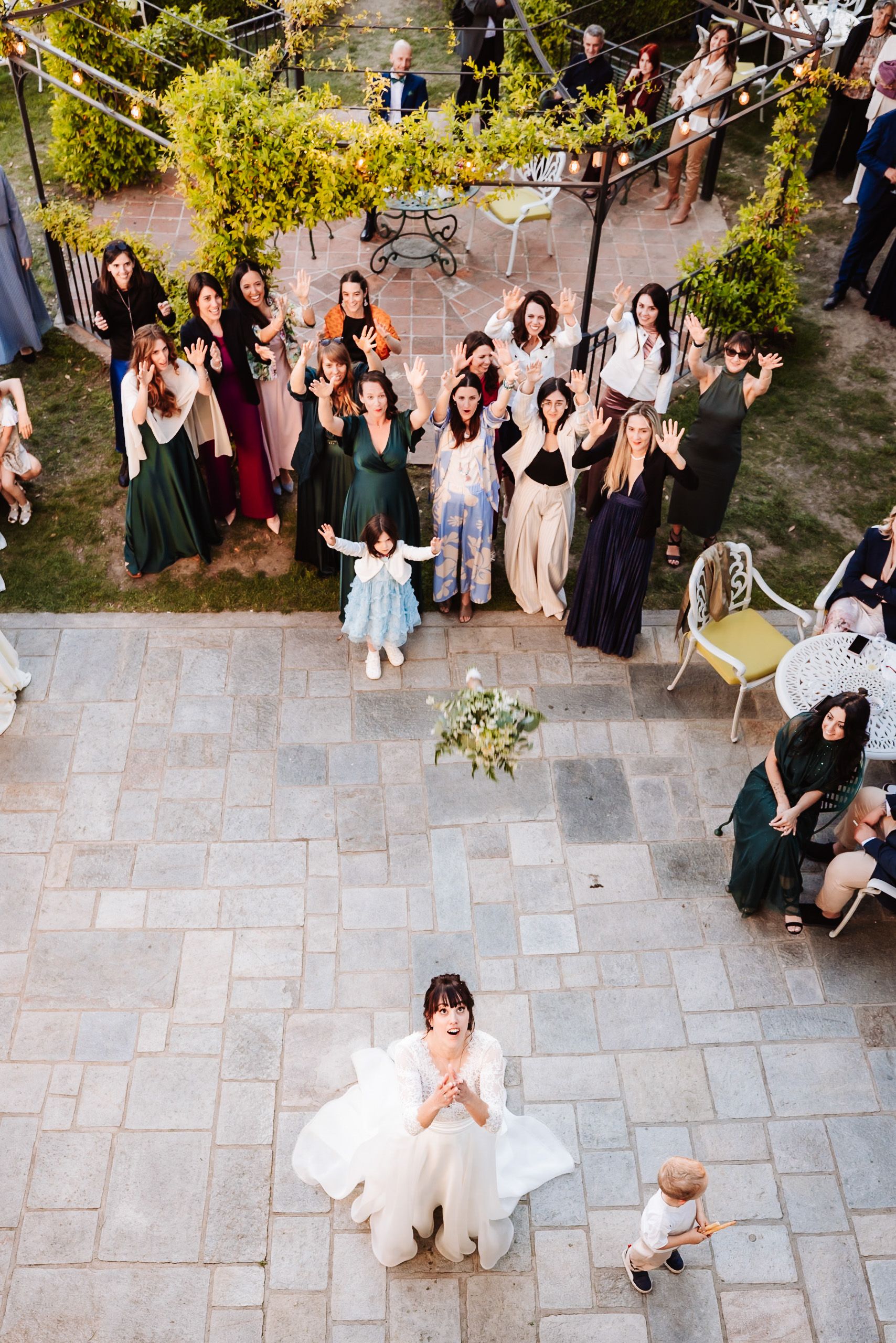 Bride tosses bouquet to excited wedding guests gathered on outdoor patio during reception celebration.