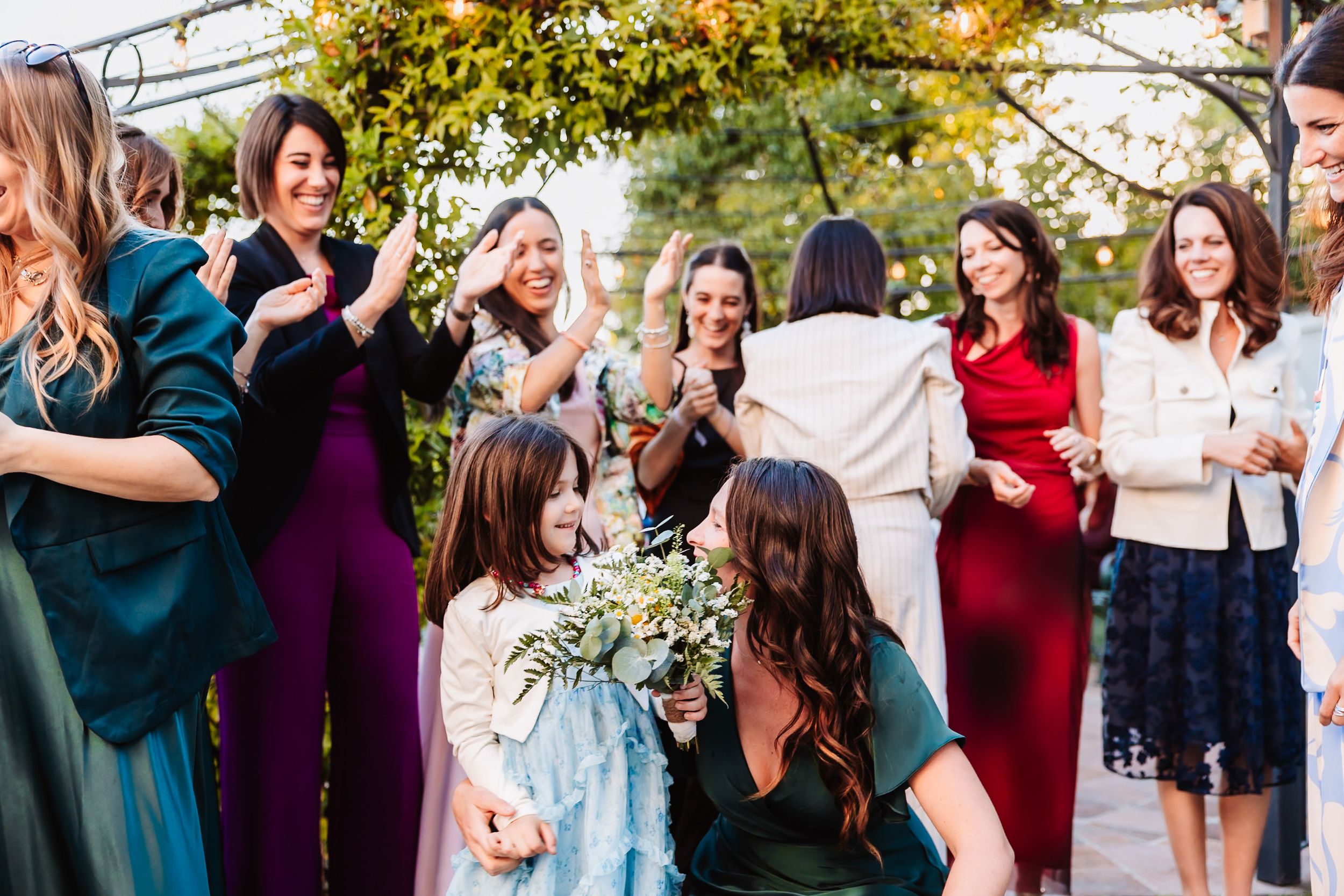 Wedding guests in colorful dresses celebrate outdoors as a woman kneels to greet a young flower girl holding a bouquet.